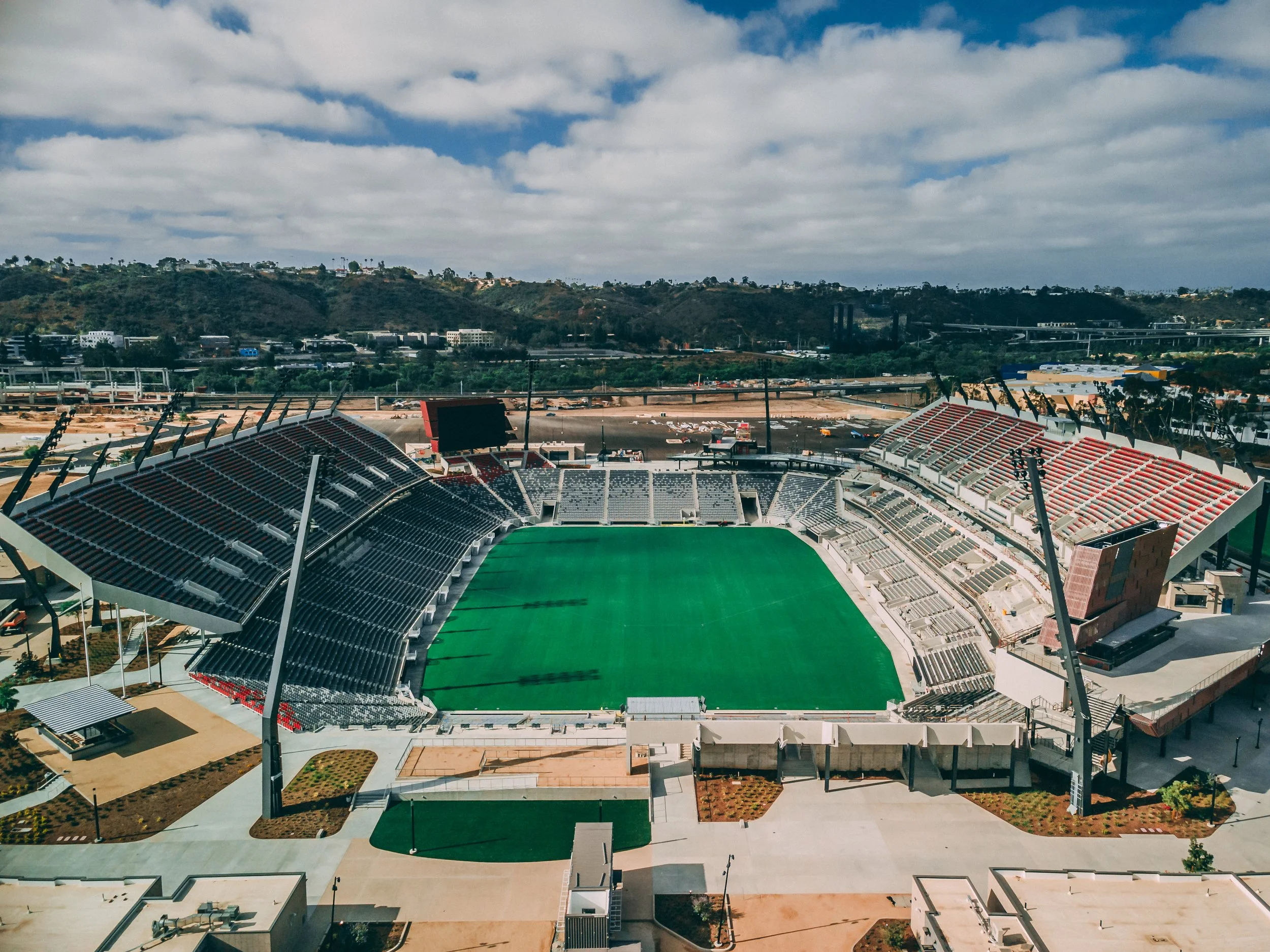 An empty Snapdragon Stadium with green field and surrounding tiered seating, under partly cloudy sky, with hills and infrastructure in background.