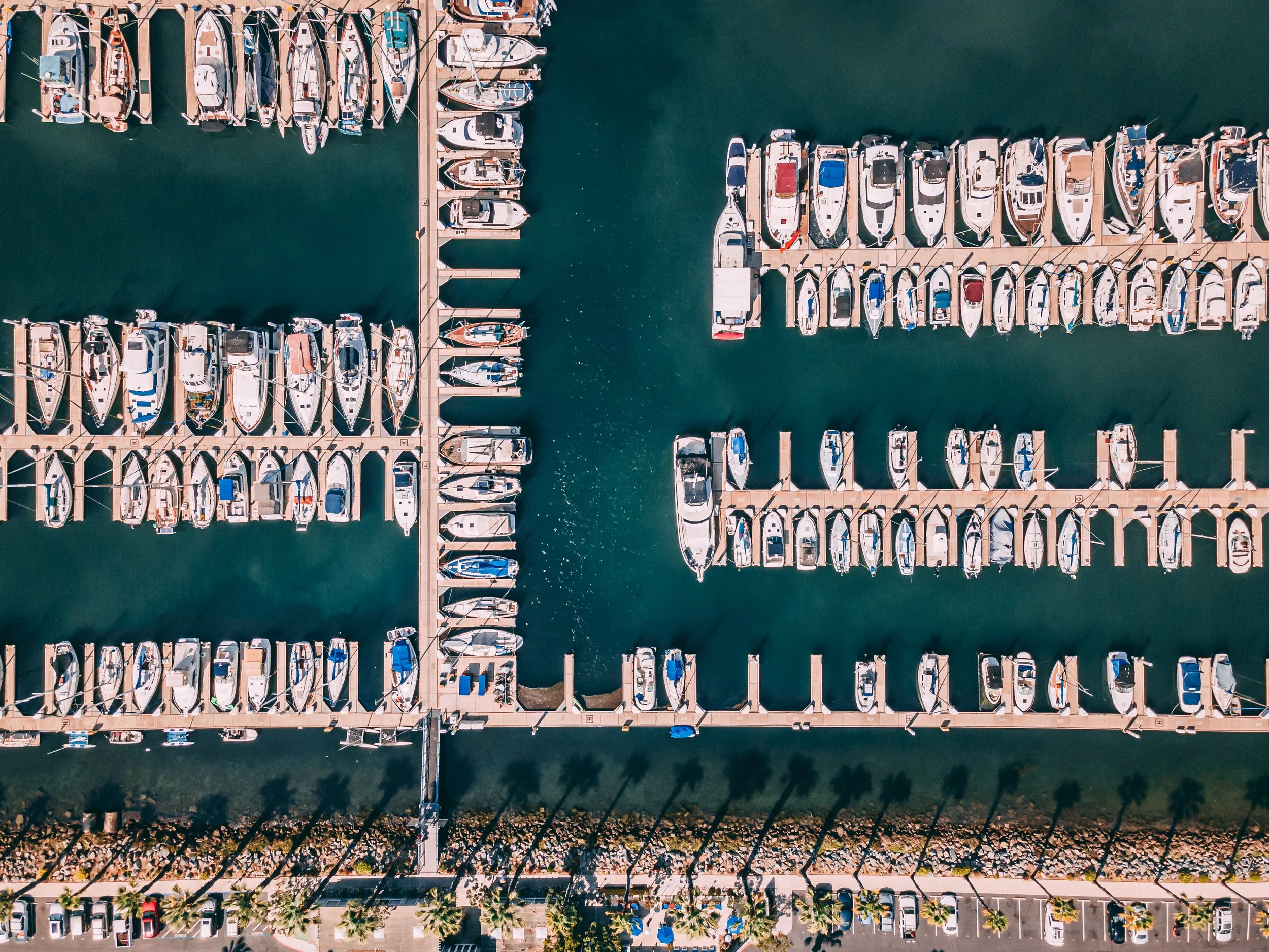 An aerial view the Chula Vista marina with multiple docks and boats, including yachts and smaller vessels, in the water, with a row of palm trees along the shoreline.