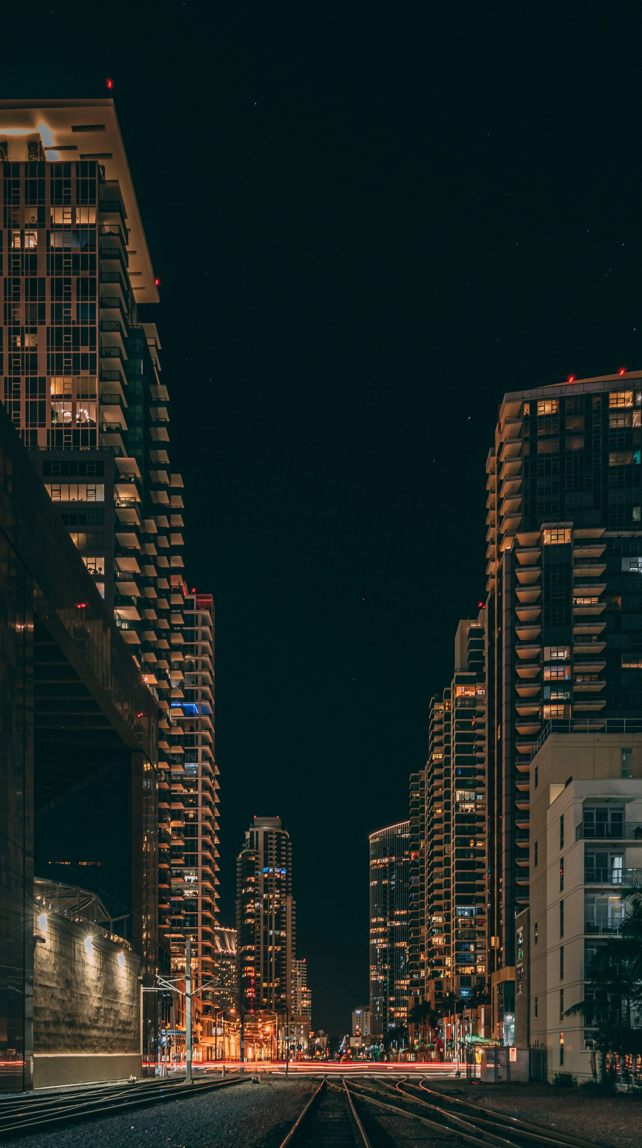 Nighttime cityscape with high-rise buildings lining a street with railway tracks, lit by streetlights and building lights.
