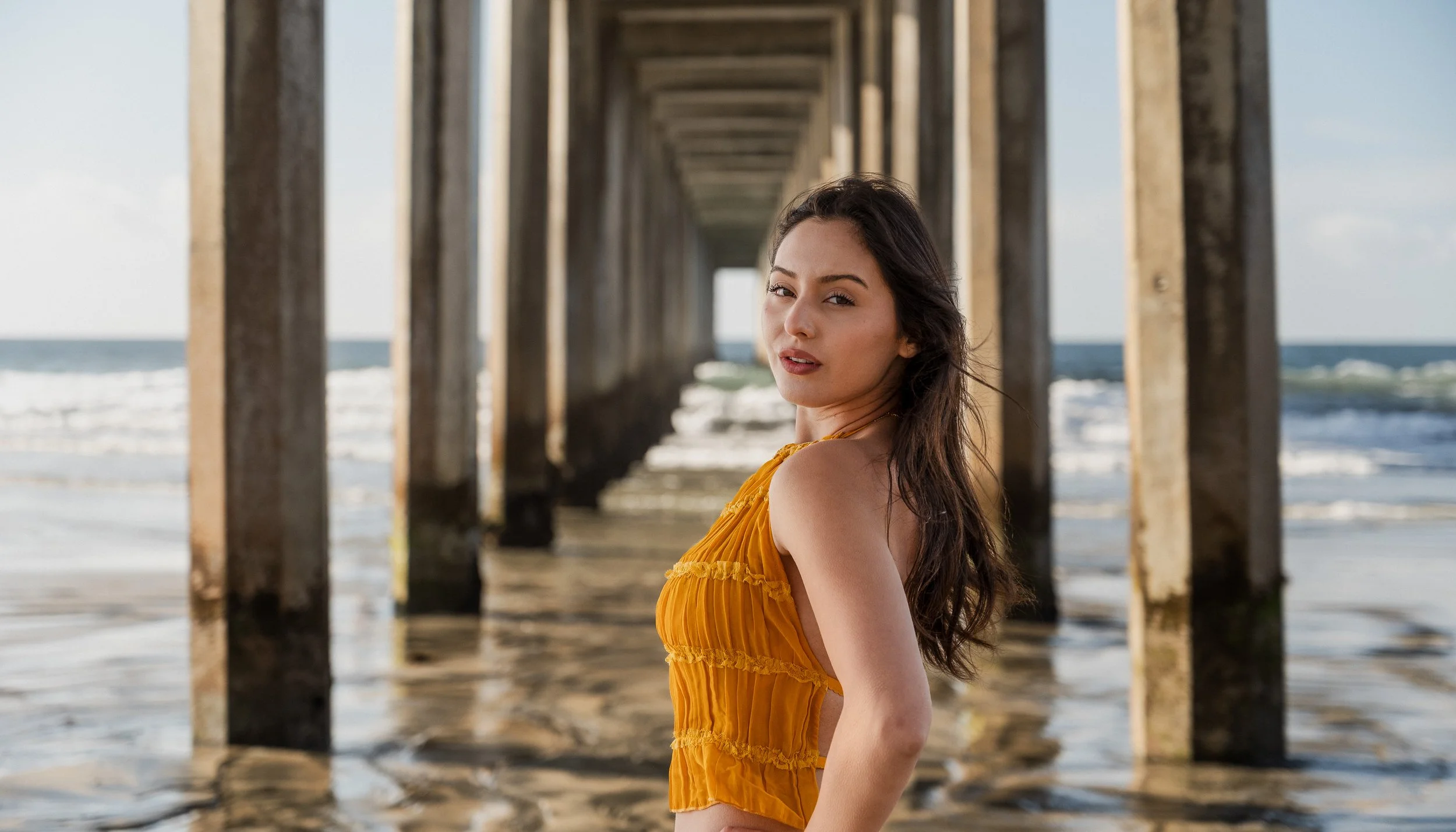 A woman in an orange dress on a sandy beach under a wooden pier, looking at the camera.