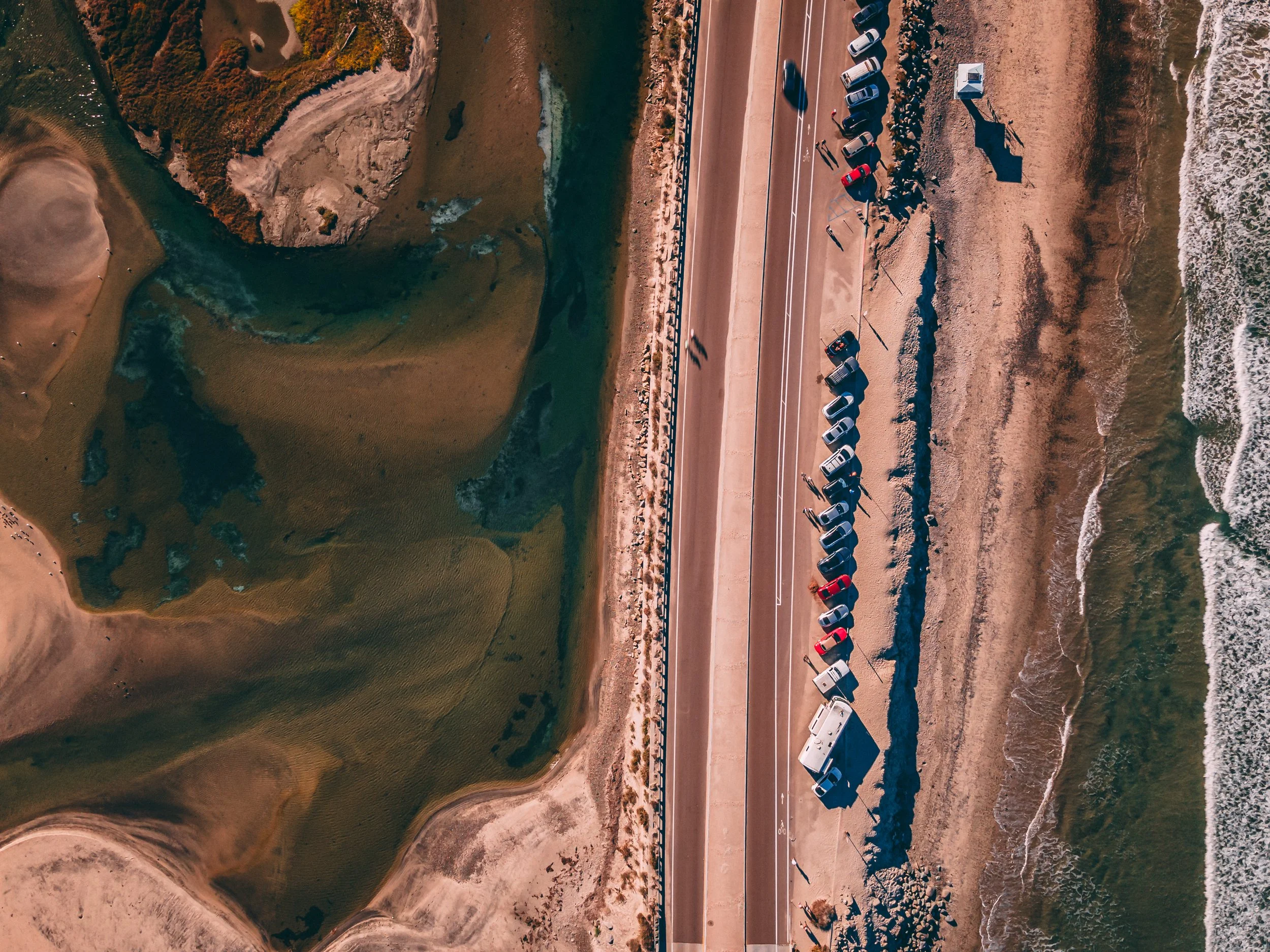 An aerial view of a coastal area with a beach, parked cars lining the road parallel to the shoreline, and a body of water on the left side with surrounding land and vegetation.