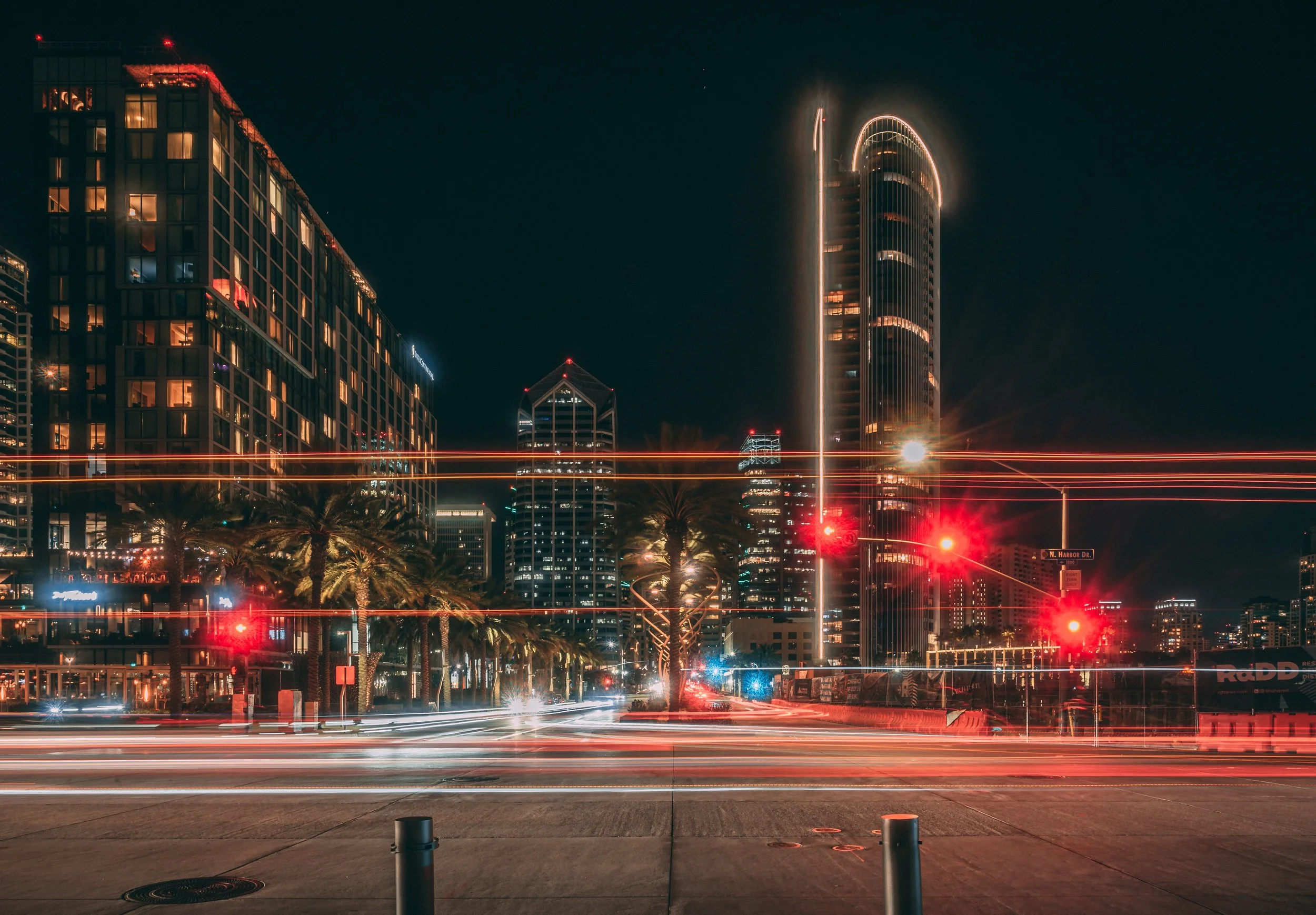 Nighttime cityscape with illuminated skyscrapers and palm trees, light streaks from moving vehicles, traffic lights, and a dark sky. At the intersection of Broadway and Harbor Drive in near downtown San Diego. 
