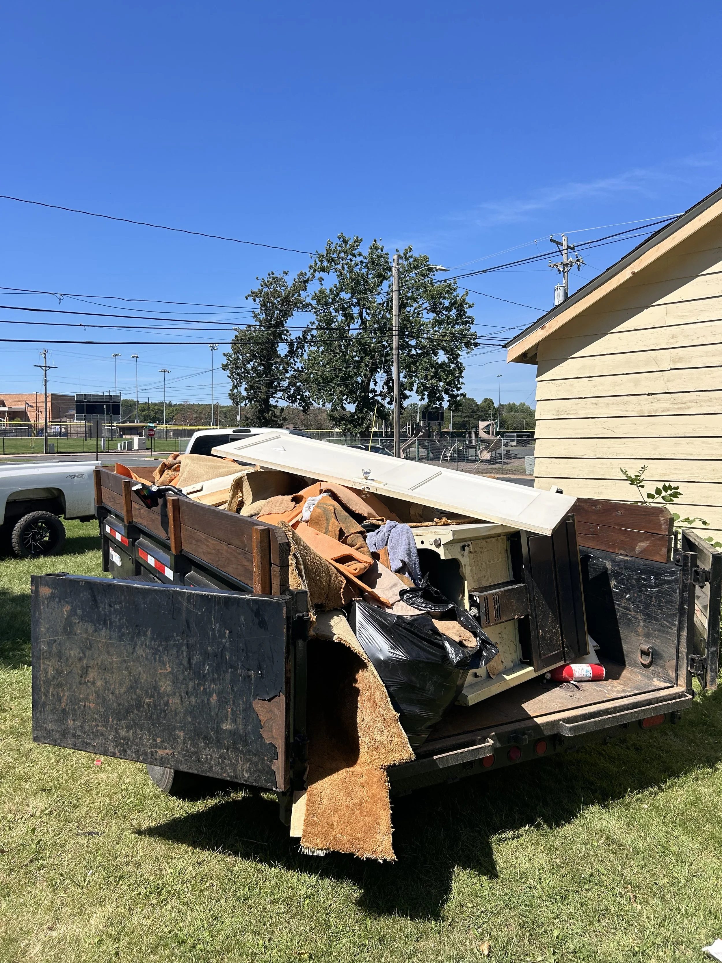 Pickup truck filled with household debris including carpets, wooden planks, and trash bags, parked on a grassy area near a building with a baseball field in the background.
