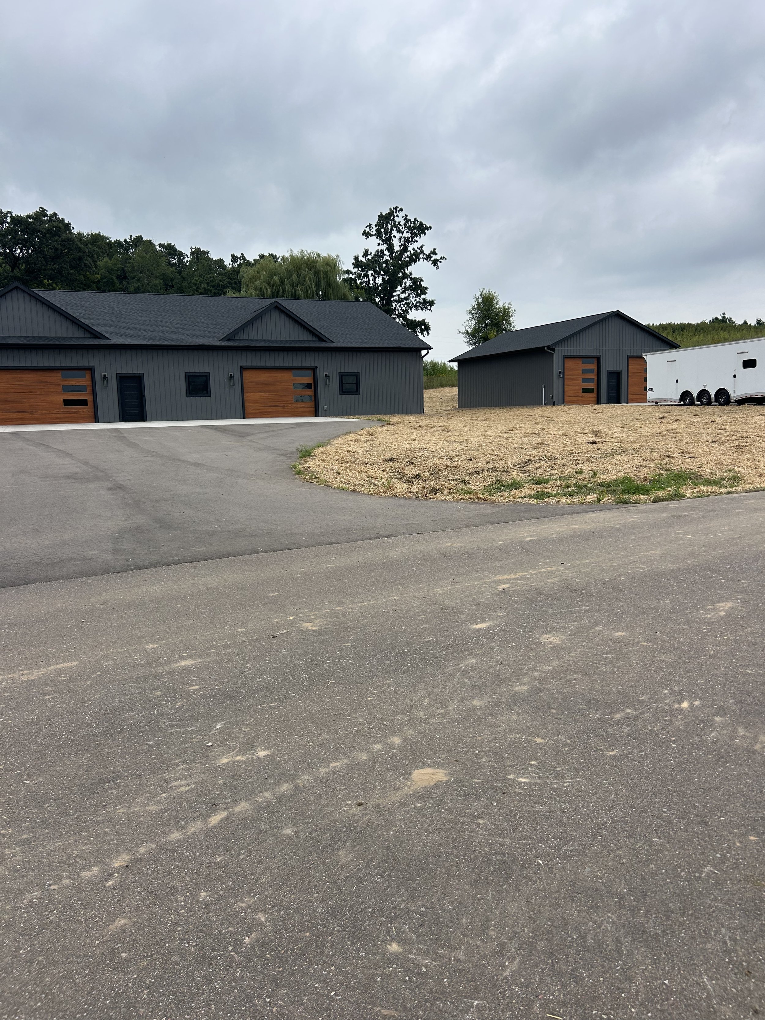 Two modern gray houses with wooden garage doors, a paved driveway, and a white trailer, under a cloudy sky, with a grassy and dirt area in the foreground.