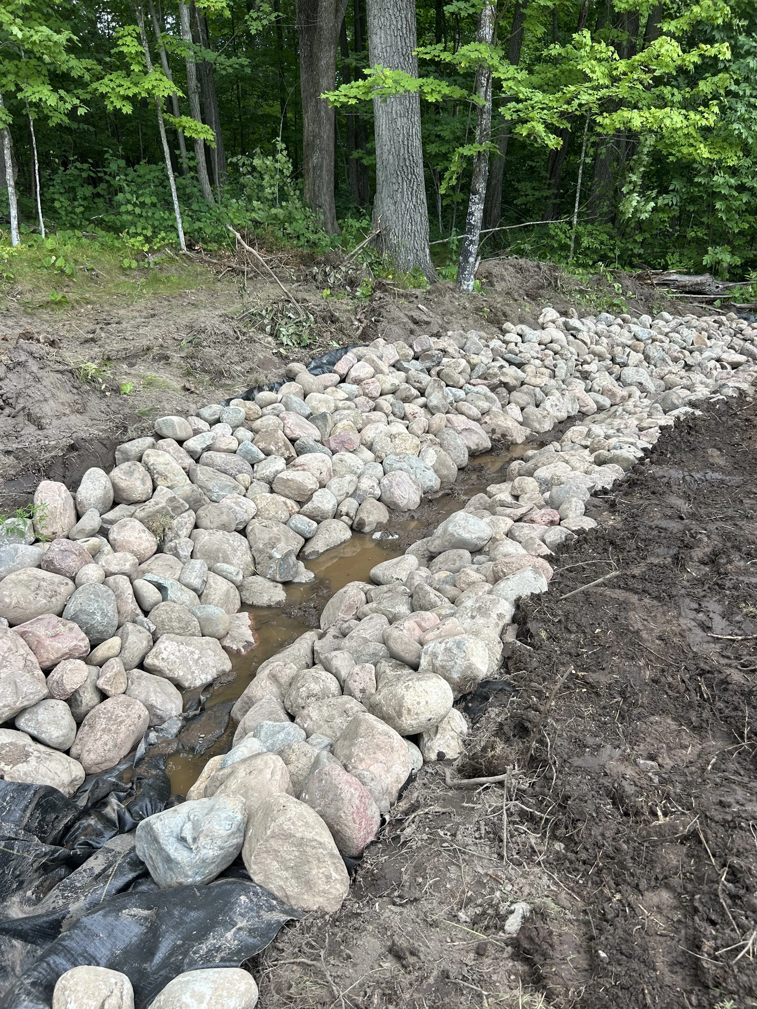 A construction site with a creek bed lined with large stones surrounded by soil and dense green trees.