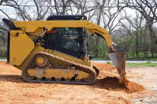 A yellow compact tracked loader grading soil at a construction site with trees in the background.