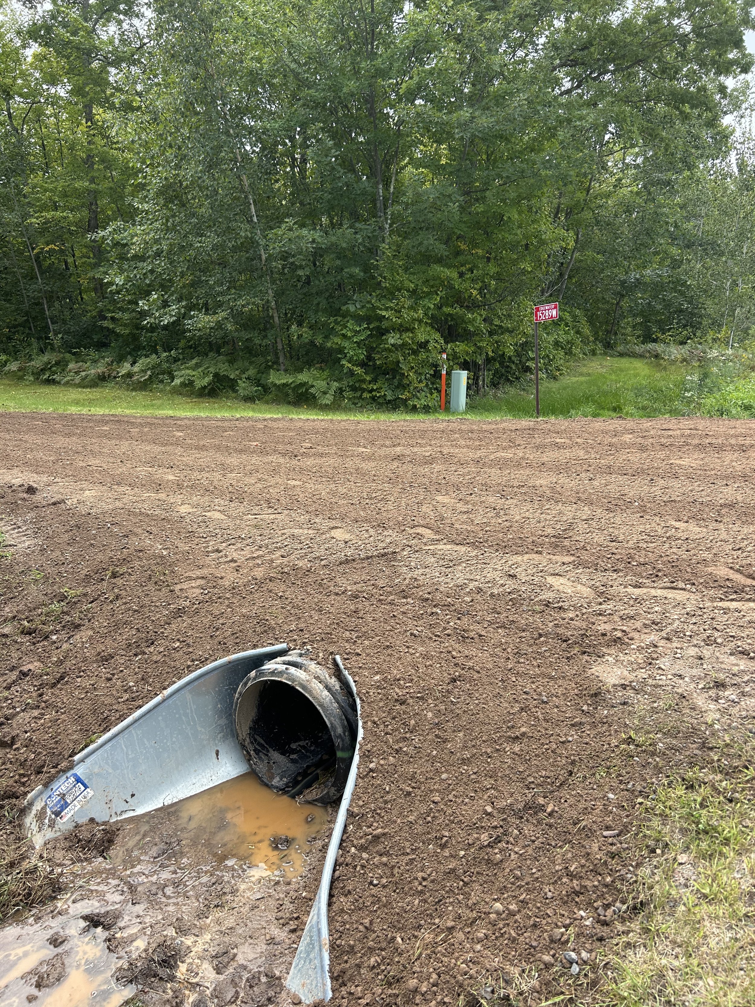 Damaged metal culvert pipe on dirt road with muddy water inside, adjacent to a grassy area with trees and a sign in the background.