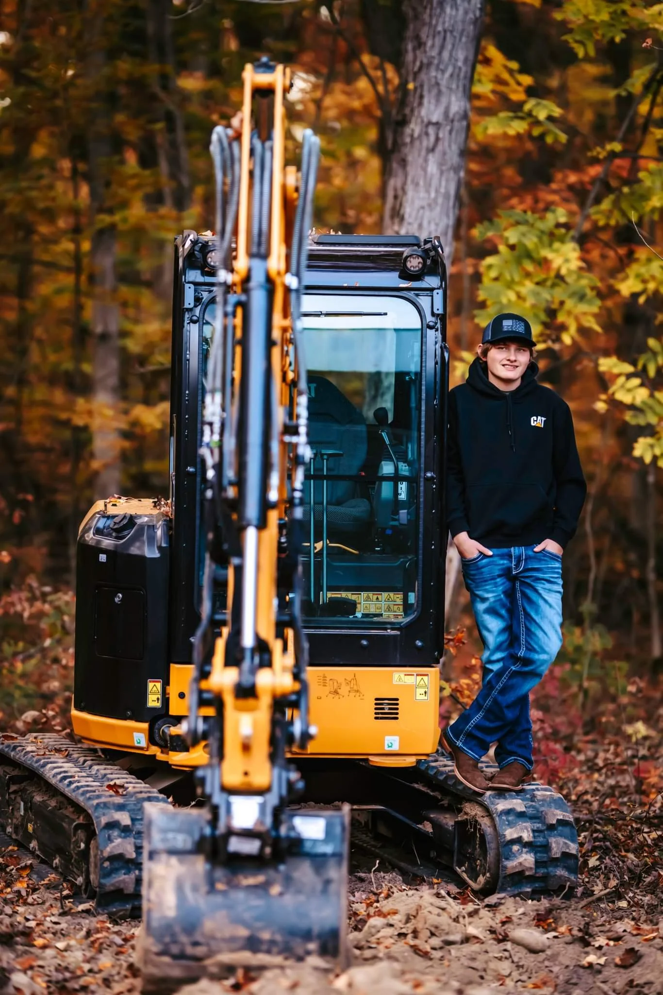 A young man in a black hoodie and jeans standing next to a mini excavator in an autumn forest with colorful leaves.