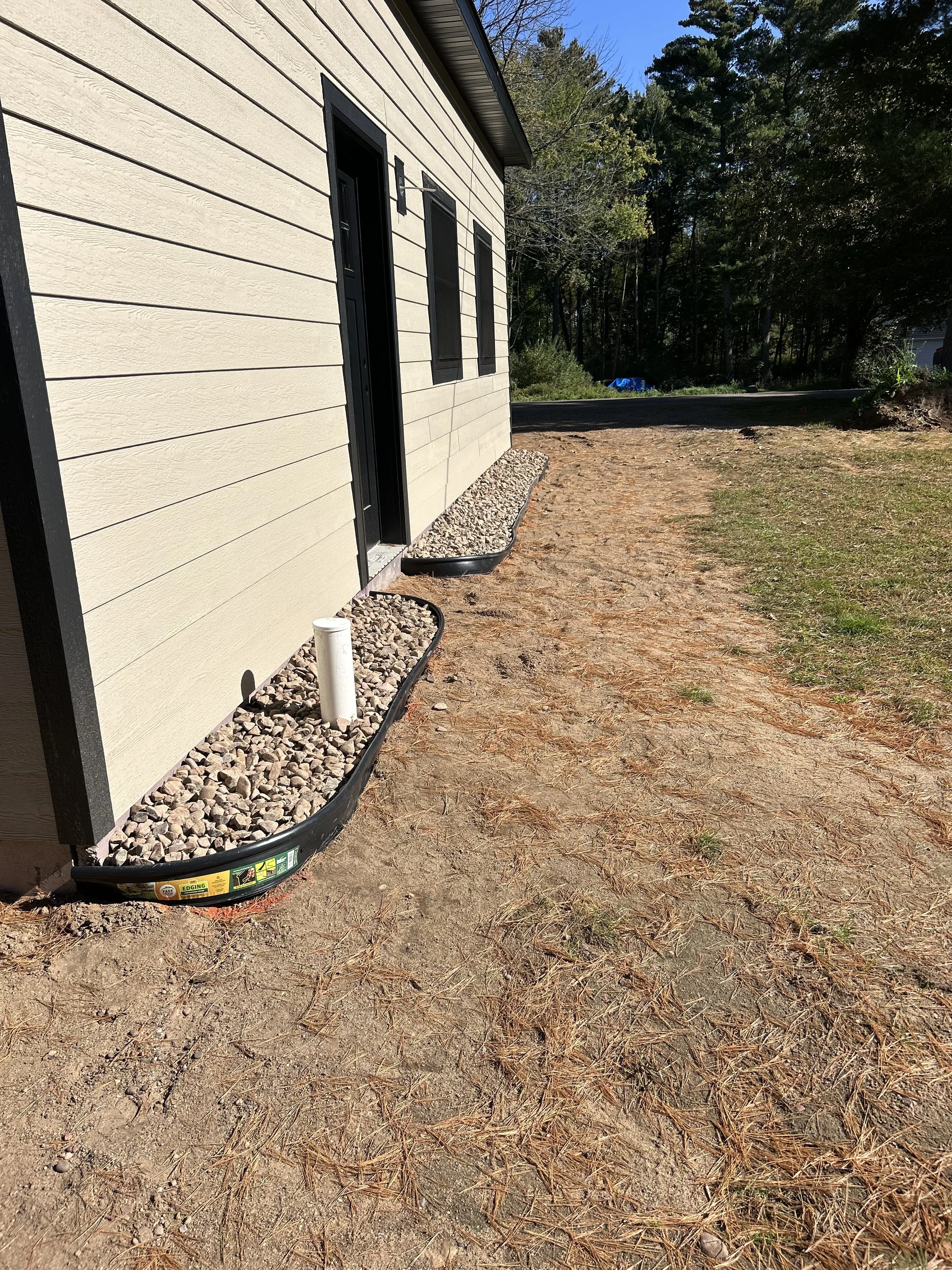 Side view of a house with beige siding, black window shutters, and a black door, with a small gravel bed underneath, and an exterior pipe protruding from the ground. The yard is partially cleared with some grass and dirt.