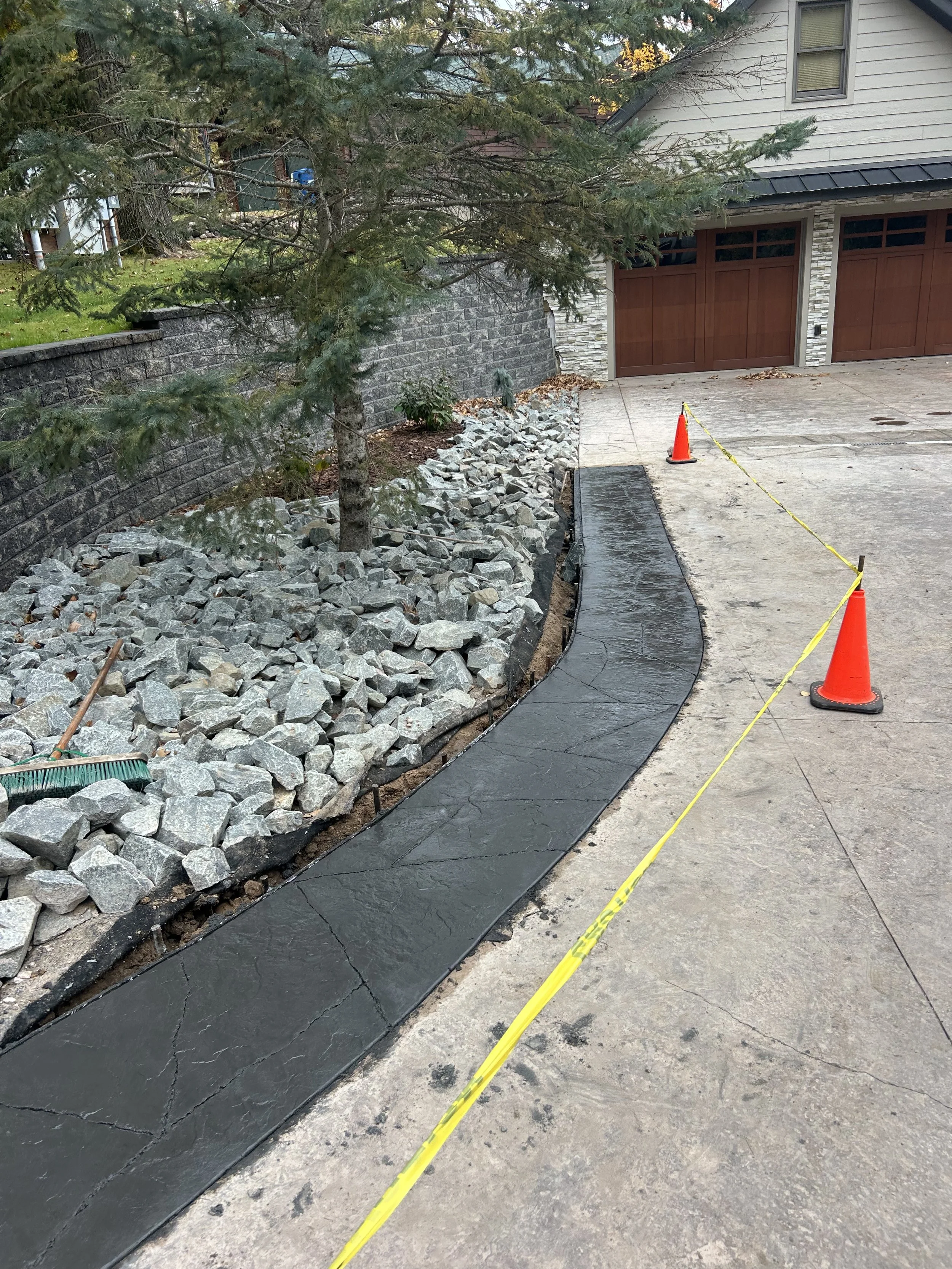 A freshly paved sidewalk with concrete and a protective barrier, flanked by a rock garden with a tree on one side and a driveway with two orange cones and yellow caution tape.