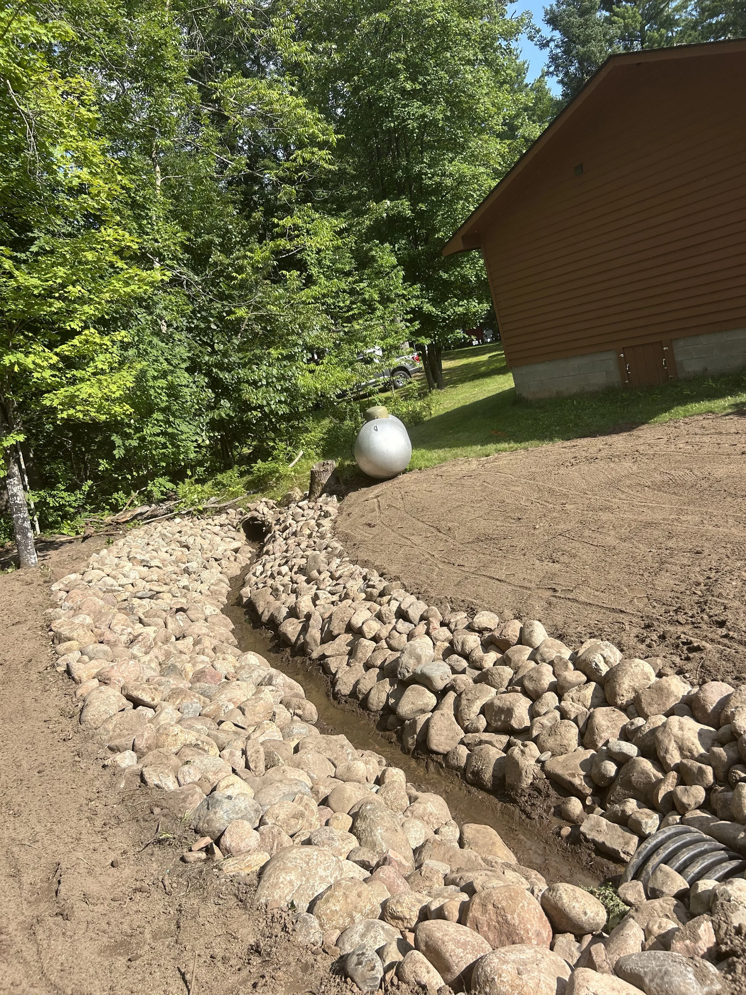 A garden bed with a stone border and a small water channel, next to a house with brown siding, surrounded by green trees on a sunny day.