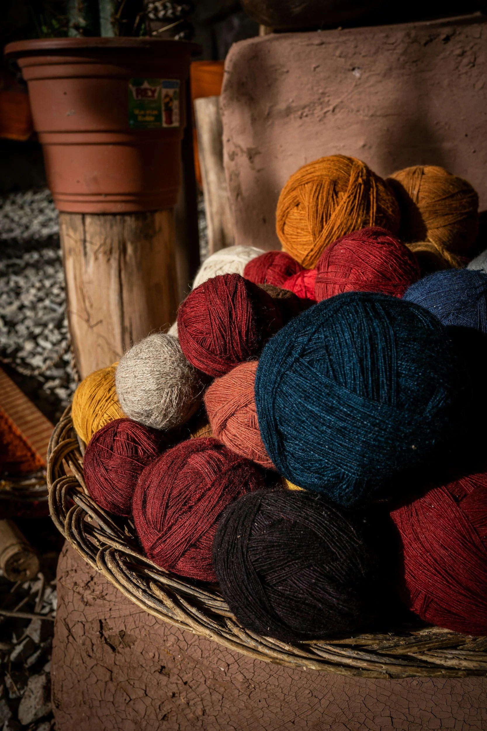A basket filled with balls of yarn in various colors including red, blue, orange, beige, and black, placed on a rustic surface with a potted plant and wooden elements in the background.