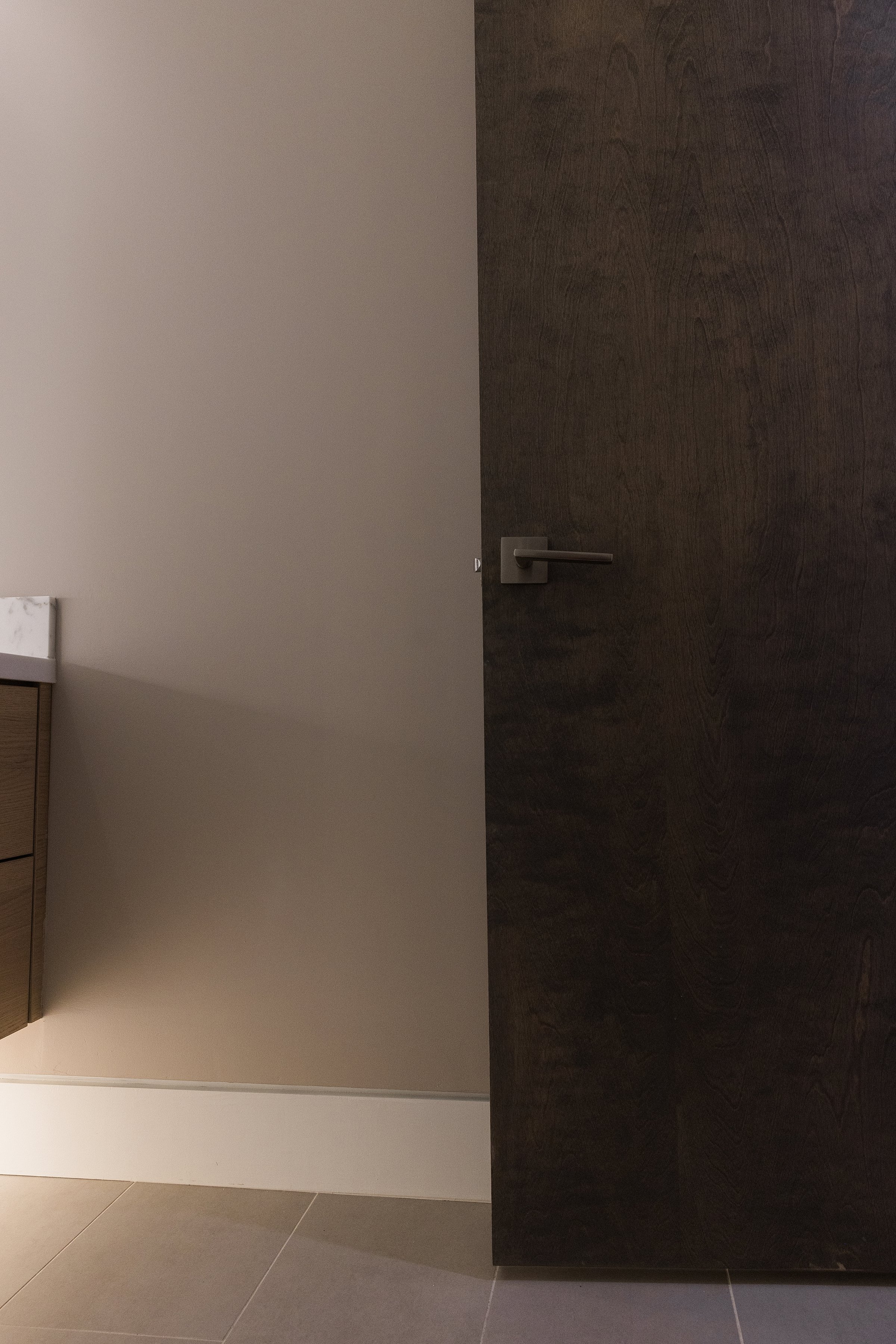 Partially open dark wooden door with a metal handle in a modern room, with beige wall and tile floor.