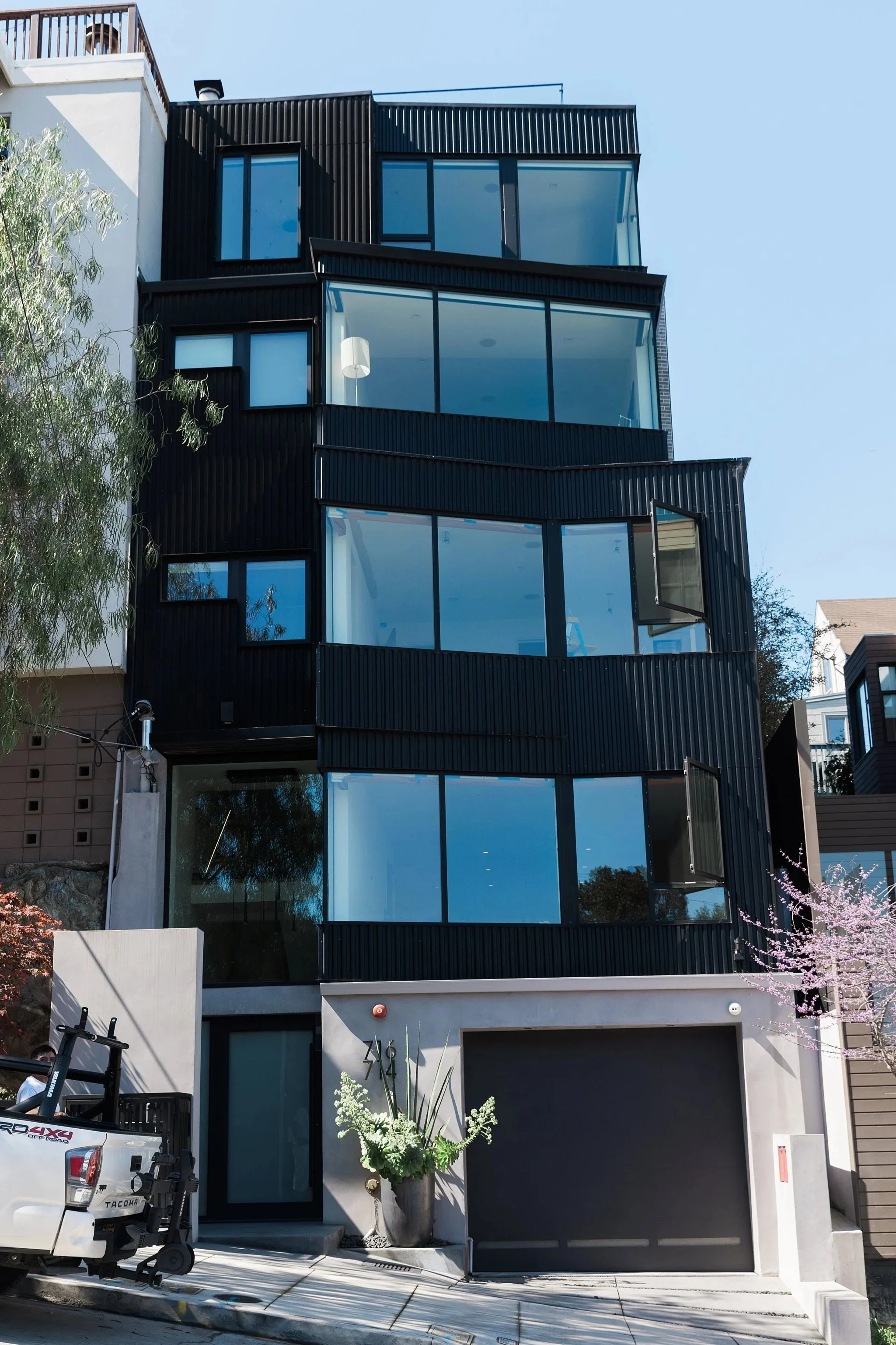A modern multi-story building with large glass windows and black exterior siding, featuring some windows open, with a tree and pink blossoms in front and a parked truck nearby.