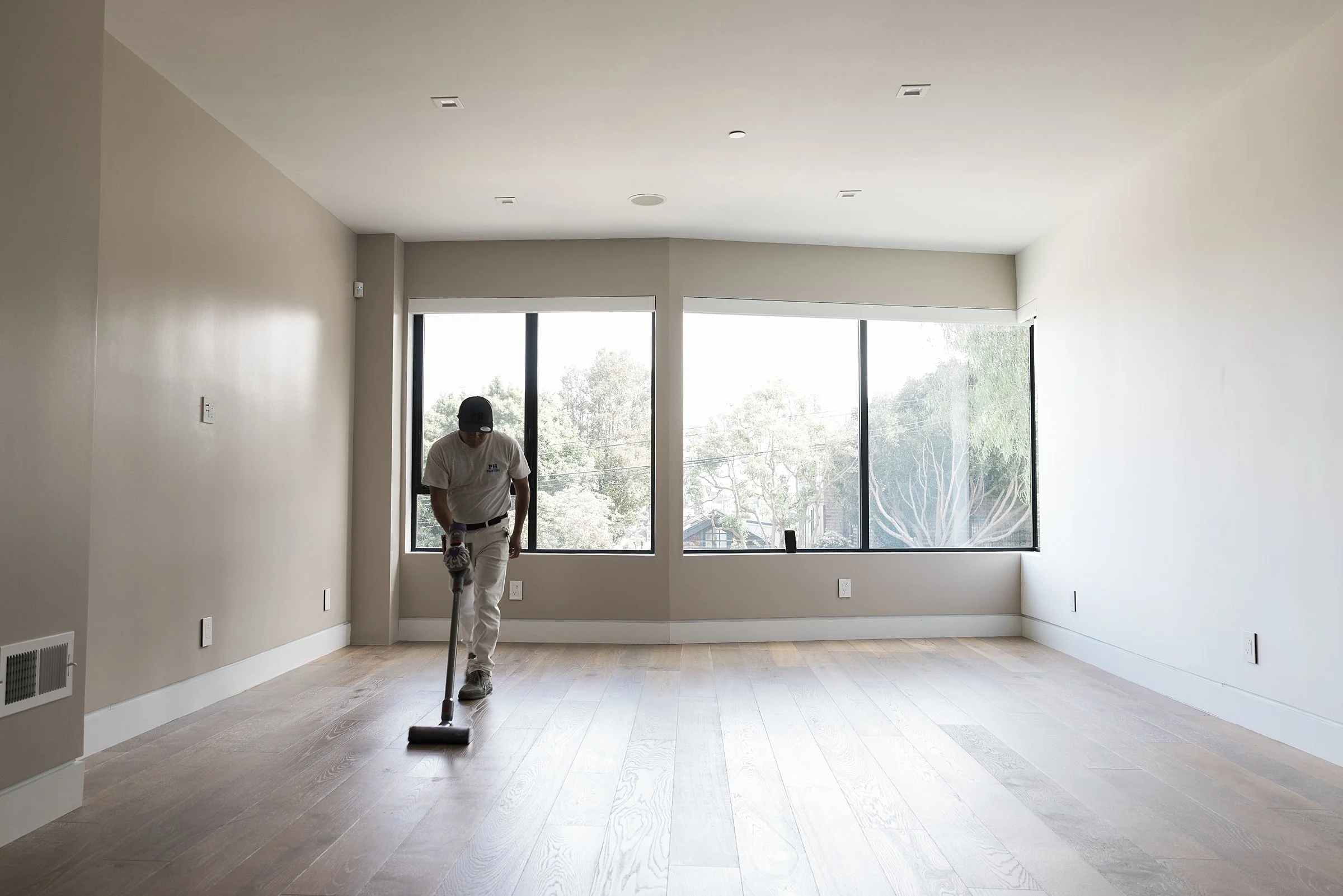 A person cleaning a spacious, empty room with large windows and light wood flooring.