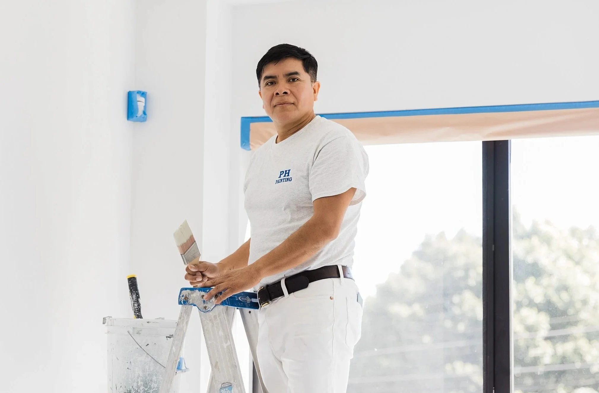 Man painting a wall while standing on a ladder in a bright room with large windows.