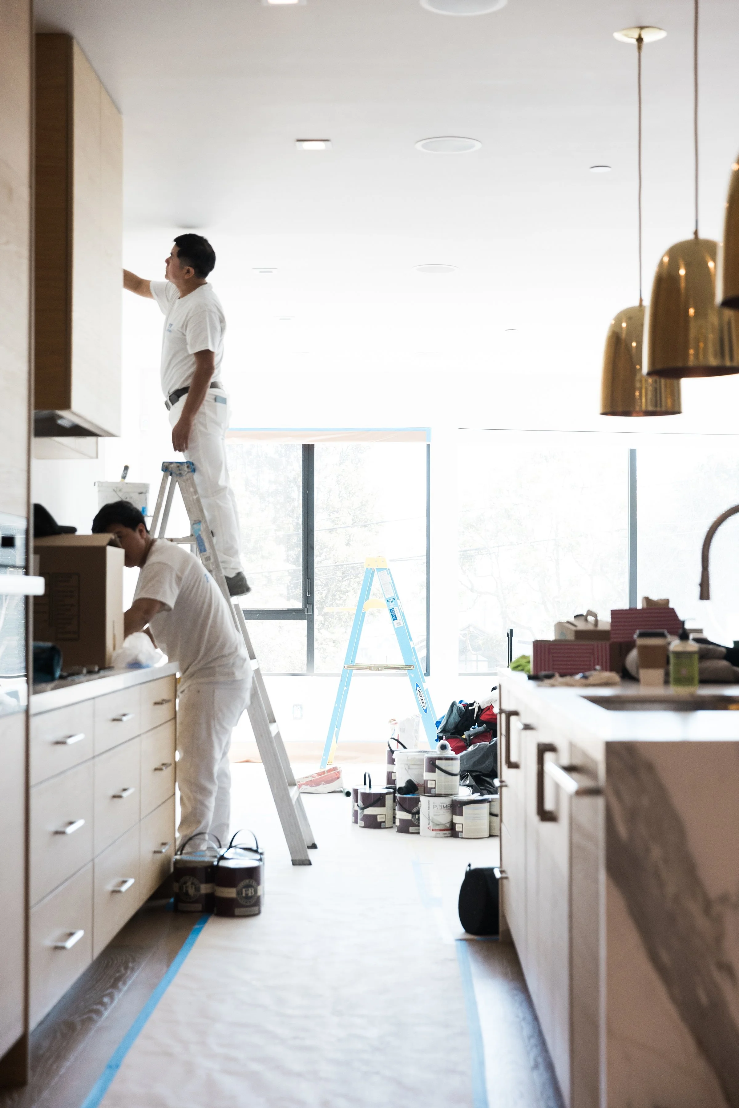 Two workers painting and preparing a kitchen area for renovation. One is on a ladder, reaching the upper cabinets, while the other is handling supplies below.