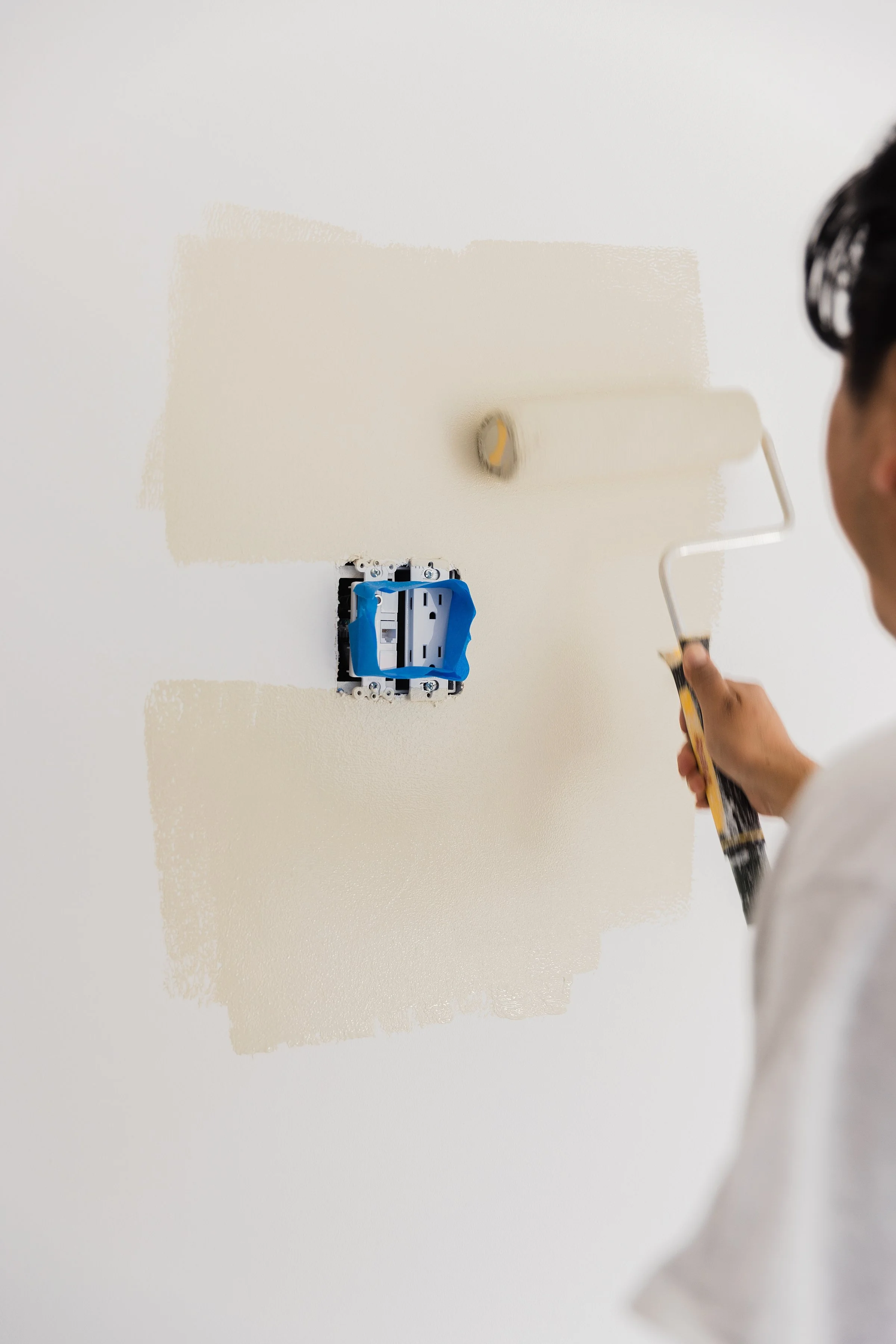 Person painting a wall with a roller brush, near an electrical outlet and a cover plate, in a room under renovation.