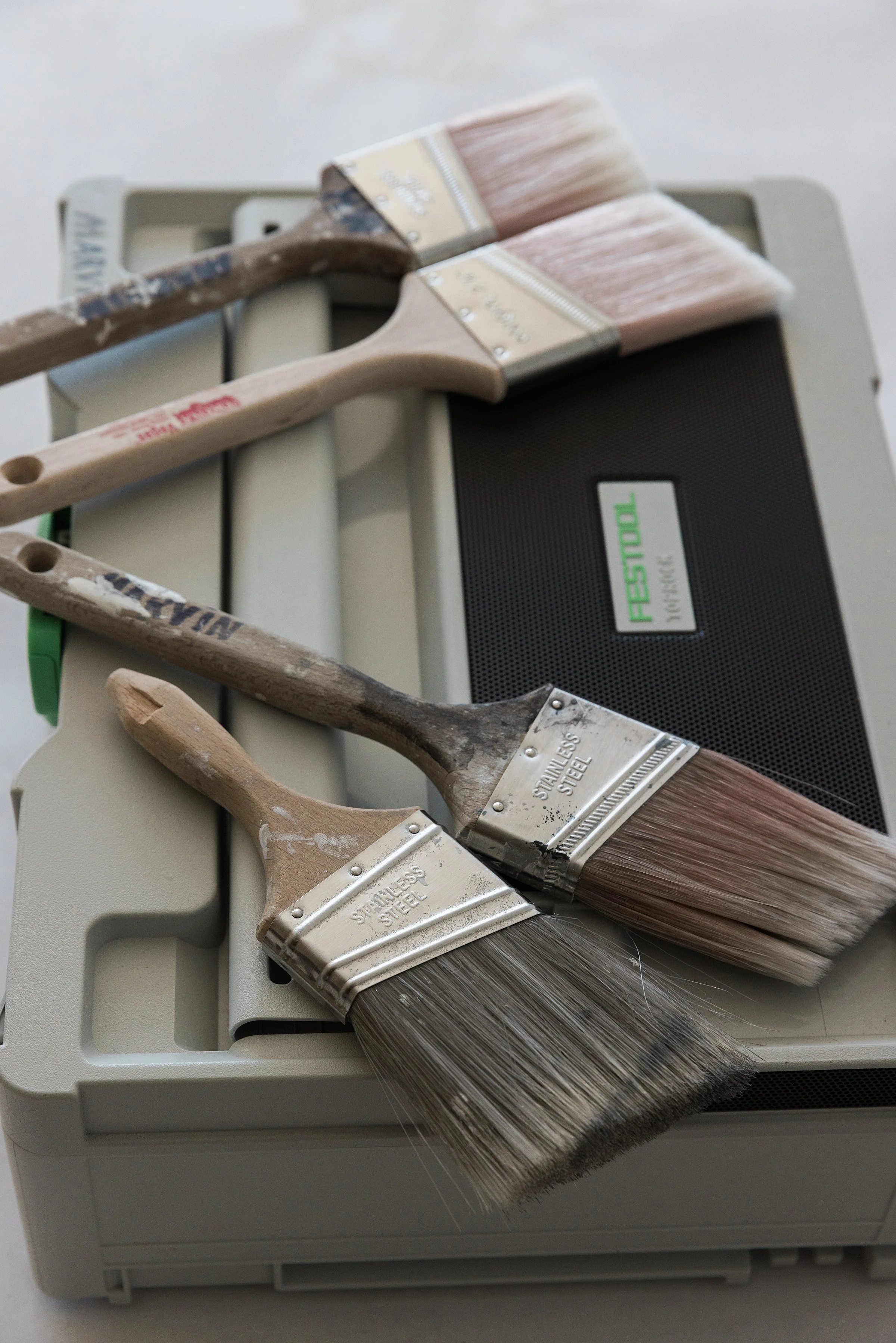 Three paintbrushes with wooden handles and bristles, placed on a plastic toolbox, with a black portable speaker nearby.