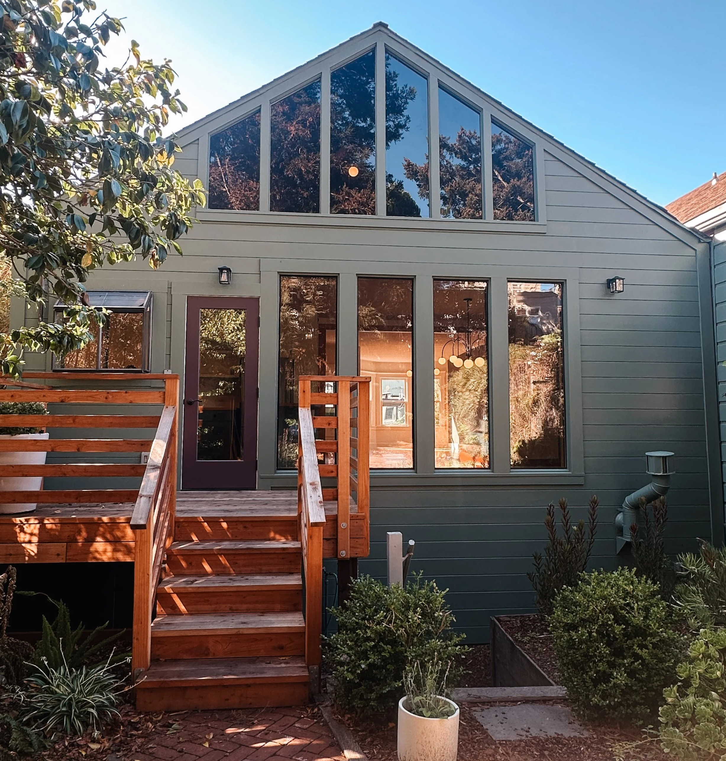 Residential house with a wooden staircase leading up to a back door and large windows, surrounded by plants and trees.