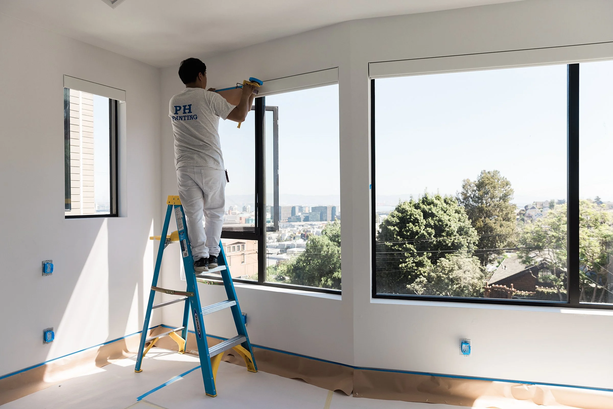 A man painting a ceiling in a room with large windows, standing on a blue ladder, with a city view outside.