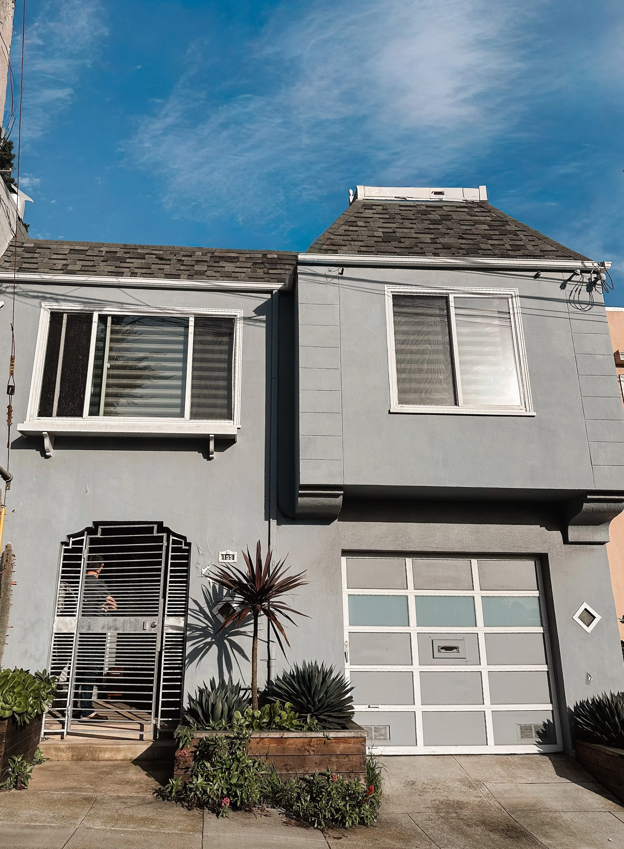 A gray two-story house with white trim, metal gate, and garage door, adorned with plants in front, under a blue sky with wispy clouds.