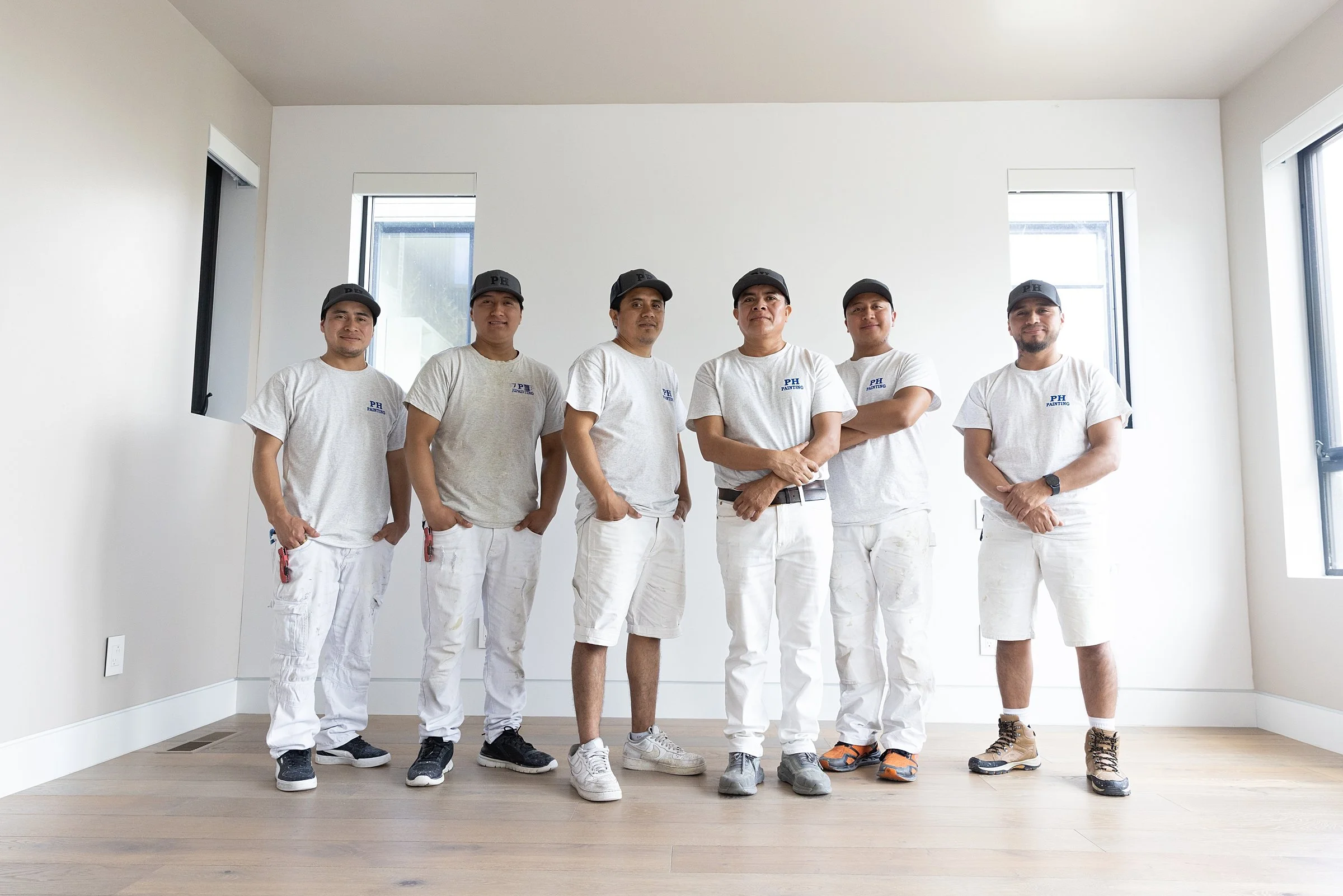Six construction workers standing inside a partly finished room with white walls and wood flooring, wearing white t-shirts, white shorts or pants, and black caps.