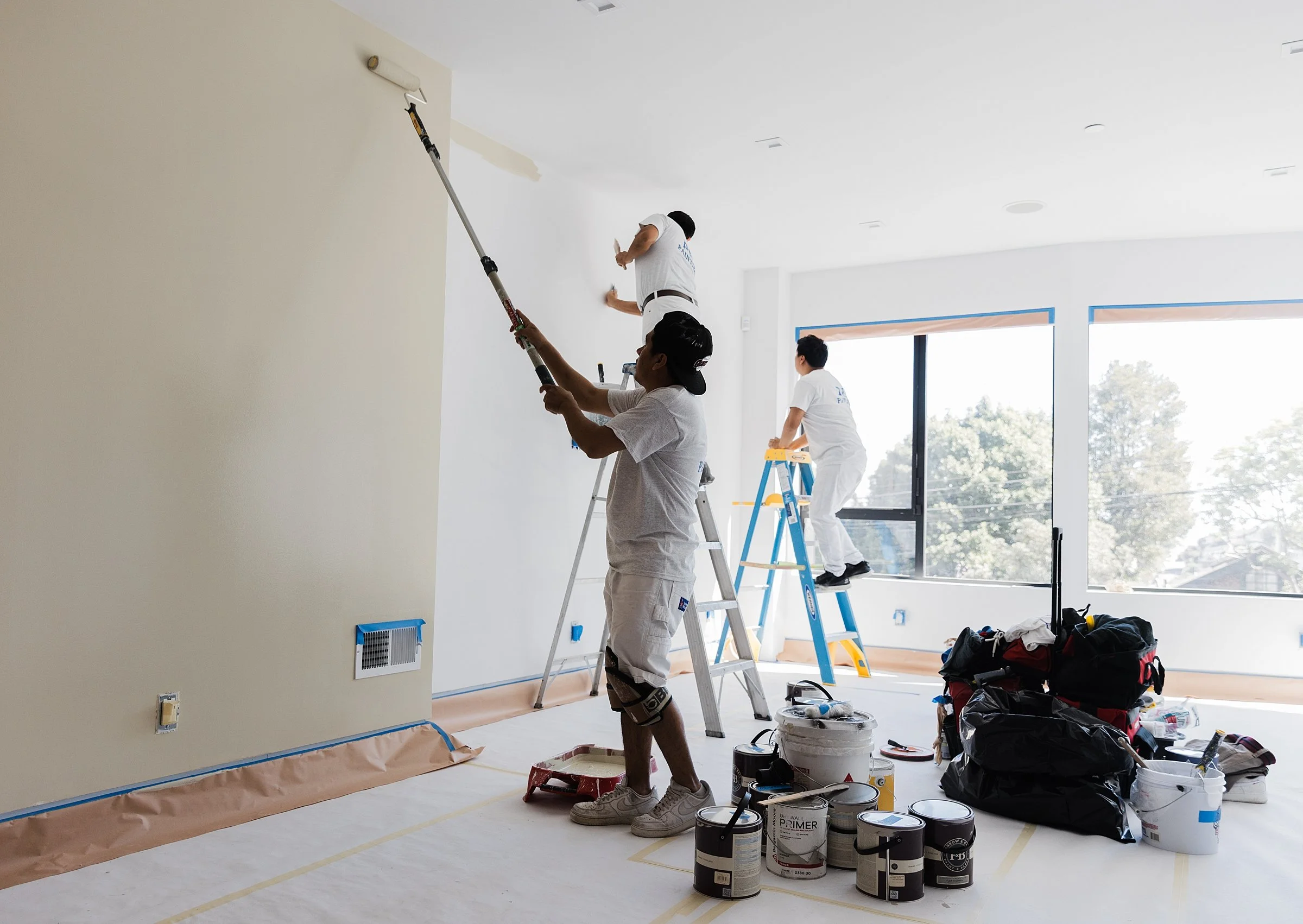 Three painters working indoors, painting a white wall with one using a roller on an extended pole, another standing on a ladder, and the third on a smaller ladder near a window. Various paint supplies and tools are scattered on the floor.