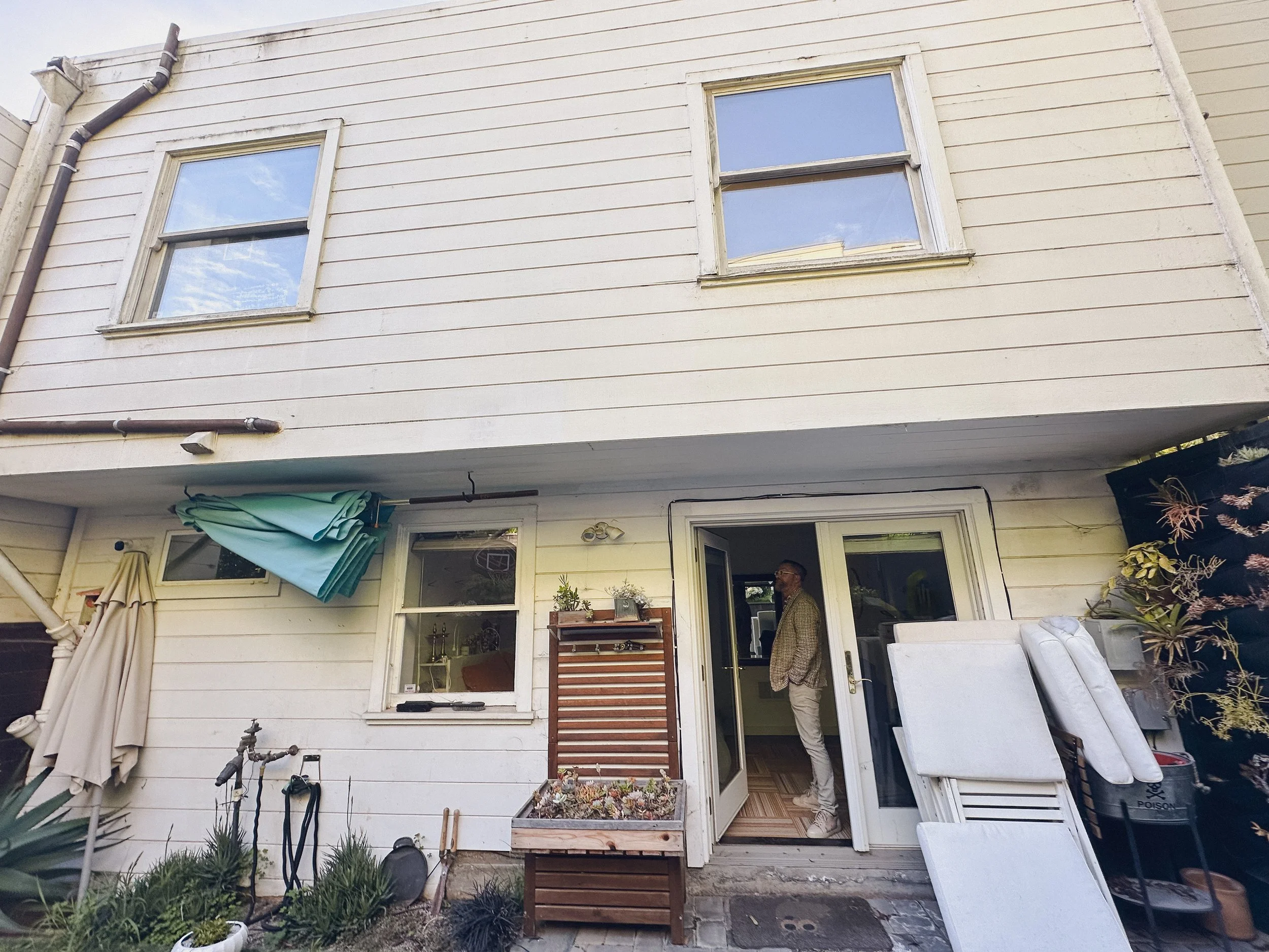 Backyard view of a two-story house with white siding, windows, and an open sliding door; a man standing inside near the doorway, outdoor furniture, plants, towels, and gardening tools.
