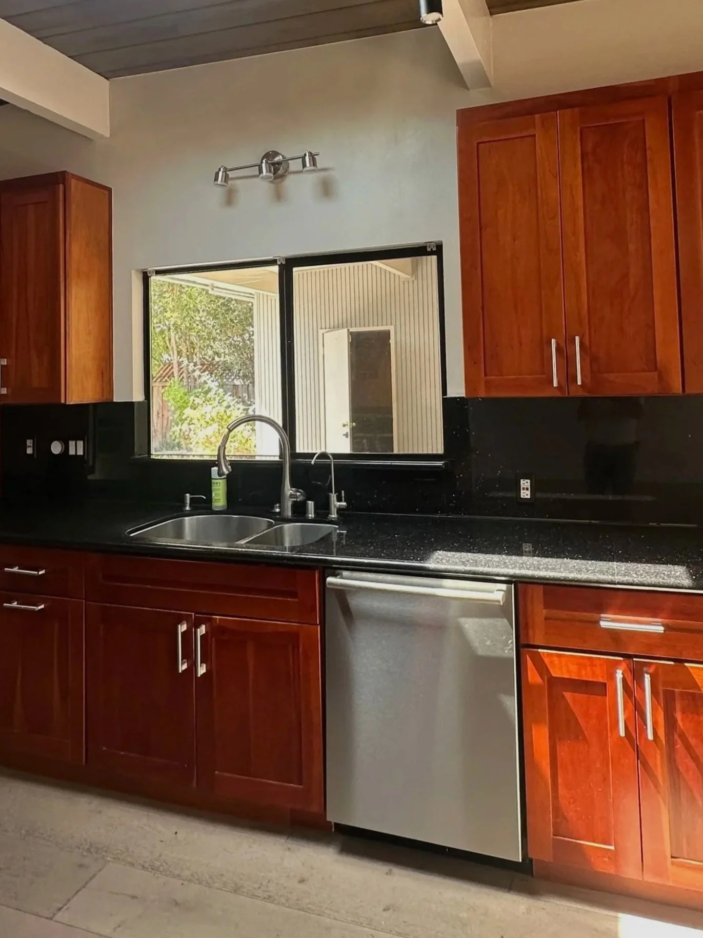 Kitchen with dark granite countertops, wooden cabinets, a double sink, and a window showing an outdoor patio with trees.