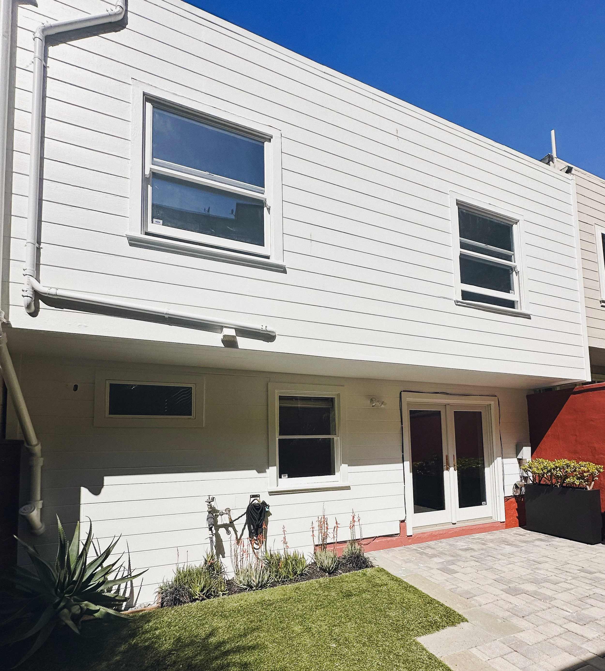 White two-story house with multiple windows, a small garden, green grass, a paved patio area, and a clear blue sky.