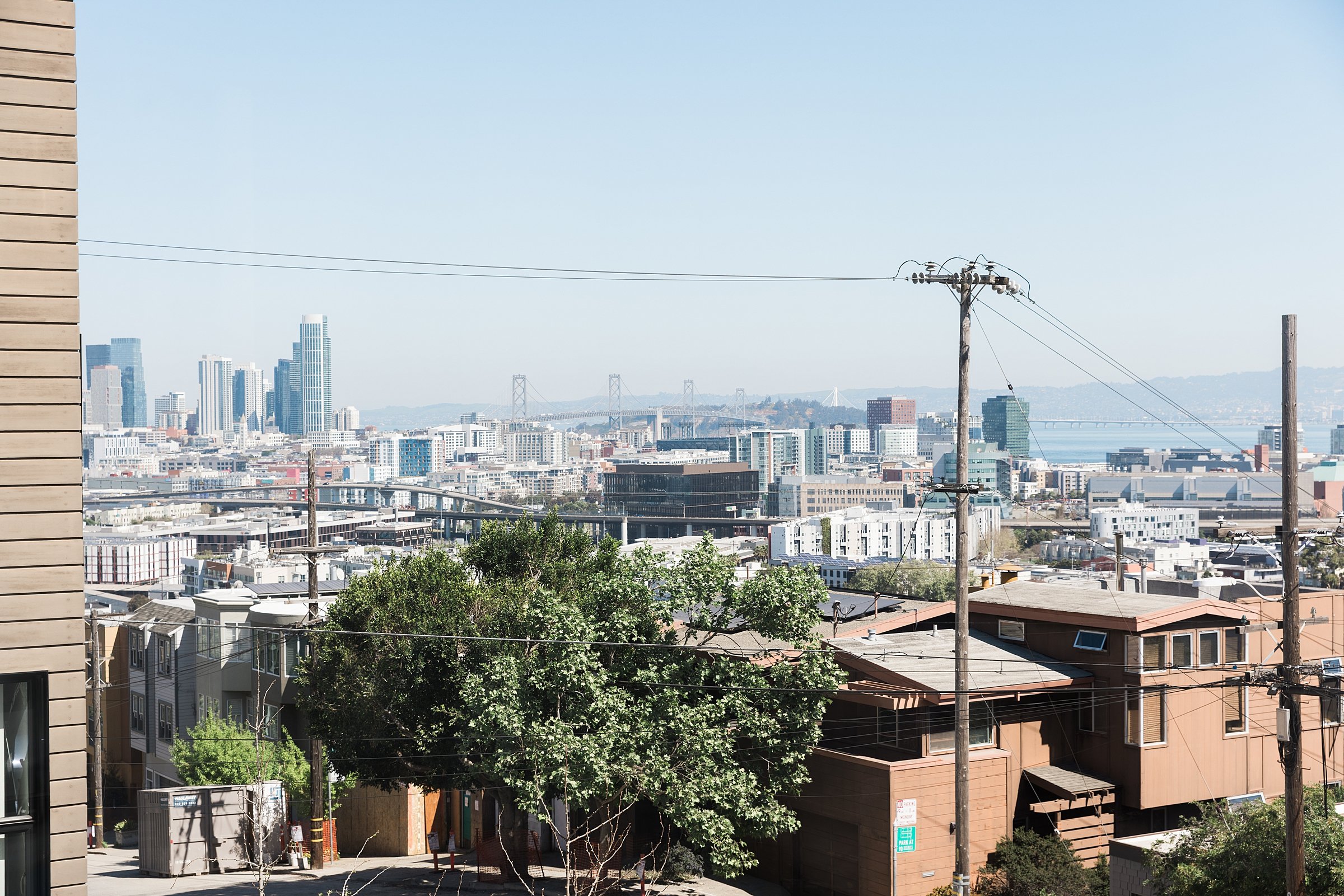 Urban cityscape with tall buildings, a bridge in the background, trees, power lines, and residential houses in the foreground.