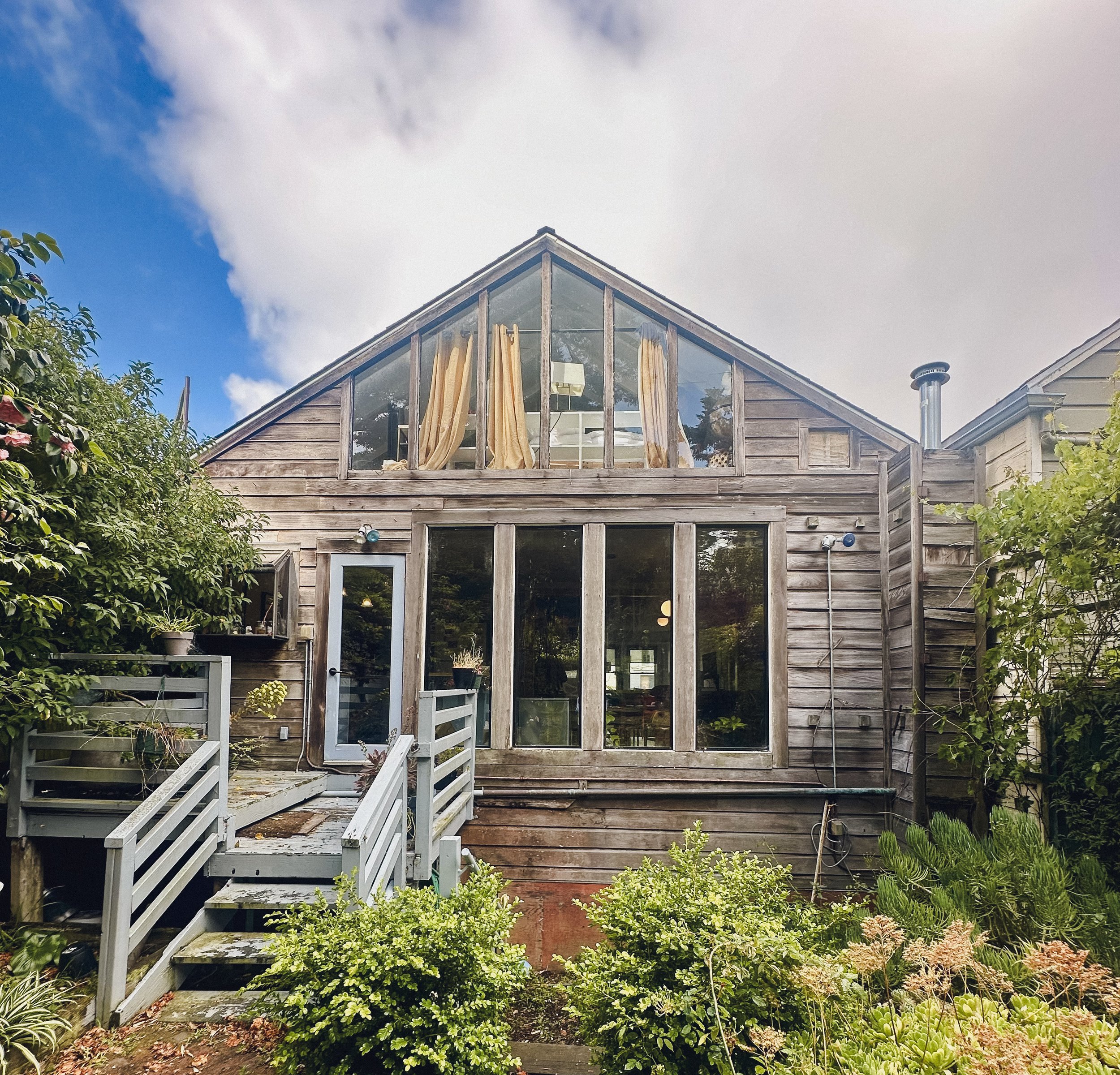 Wooden house with large glass windows and a small porch surrounded by green plants and trees, under a partly cloudy sky.