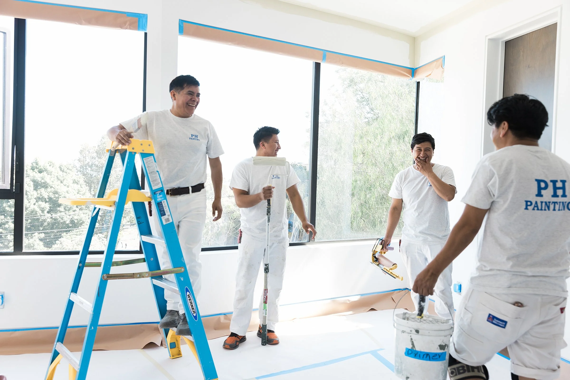 Five painters in white uniforms decorating a large window with masking tape and paper, smiling and laughing. One is on a ladder, and another is holding a paint roller. The room is bright with sunlight.