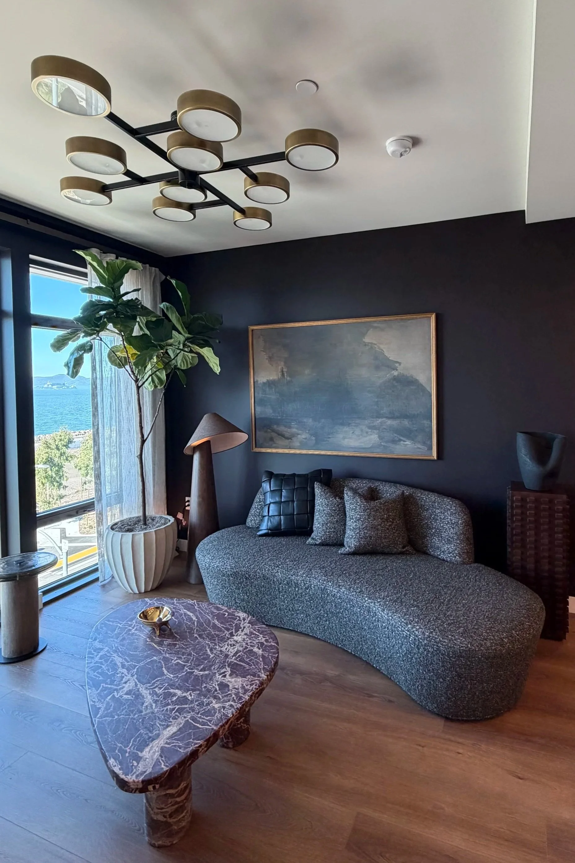 Modern living room with a curved chaise lounge, marble-top coffee table, potted plant, large window with curtains, abstract artwork, and decorative sculpture.