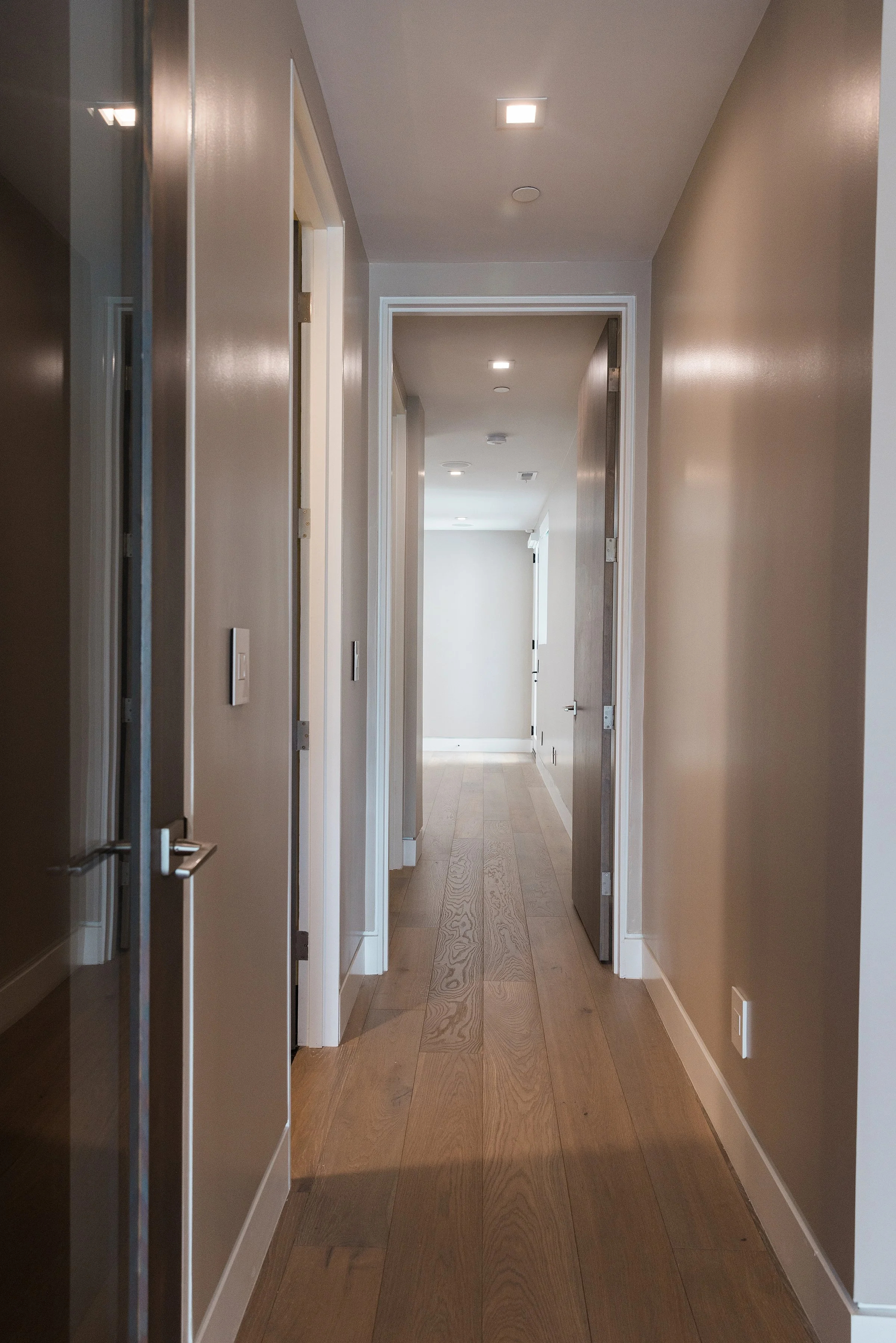 Empty hallway in a modern home with beige walls, wooden flooring, and multiple doors leading to different rooms.
