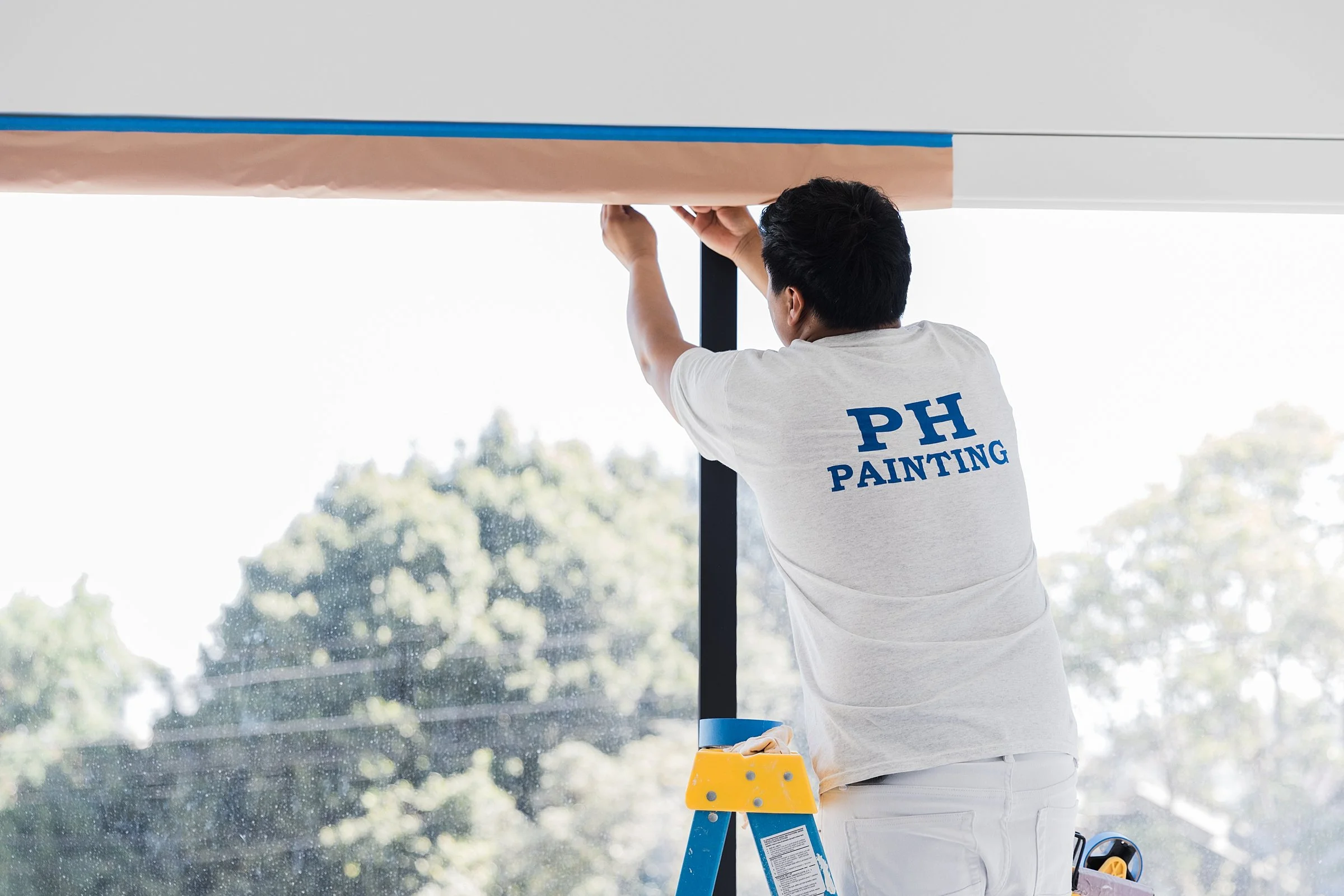 A handyman on a ladder installing or repairing a ceiling or fixture near a window.