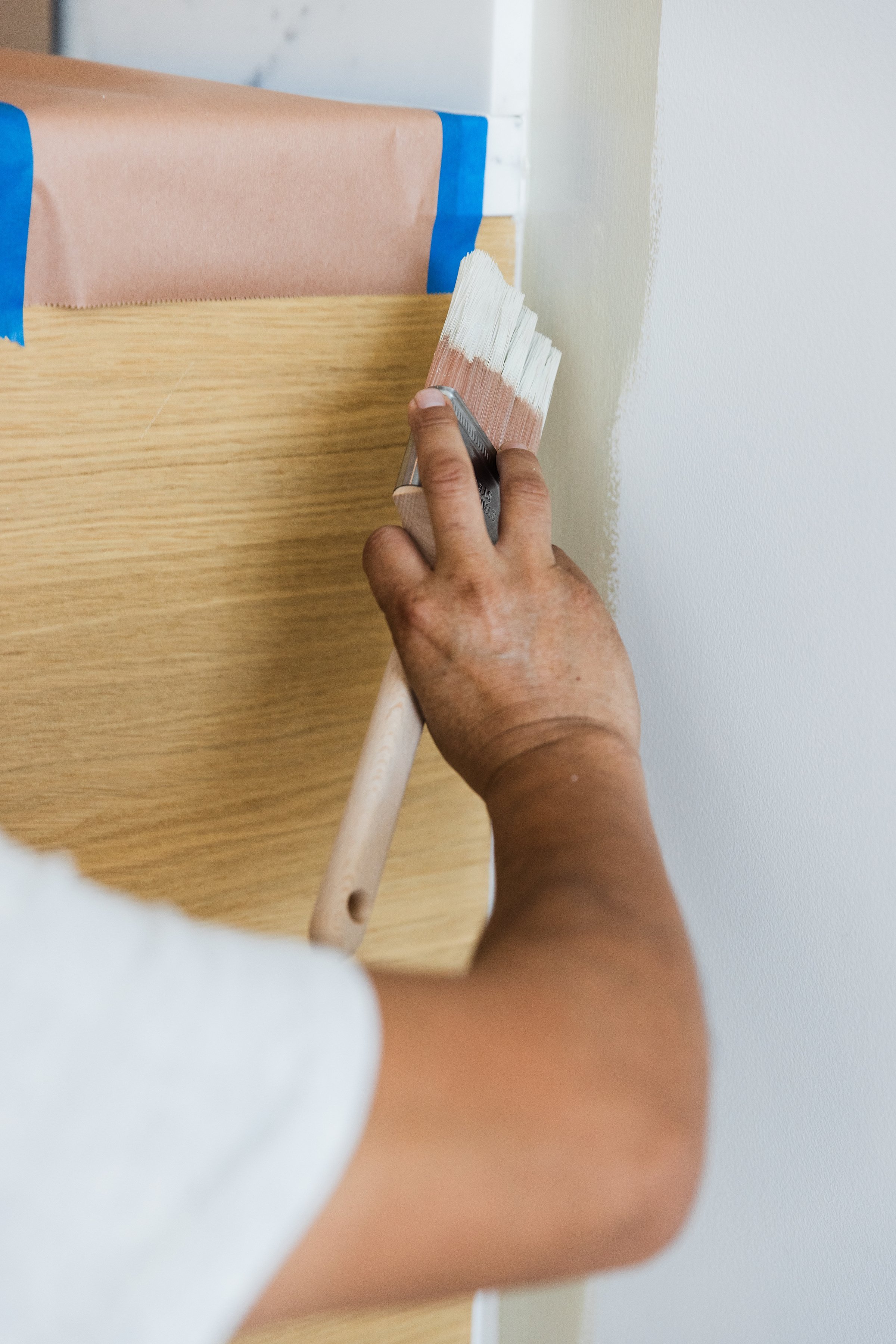 Person painting a wall with a paintbrush, next to a piece of furniture covered with painter's tape for protection.