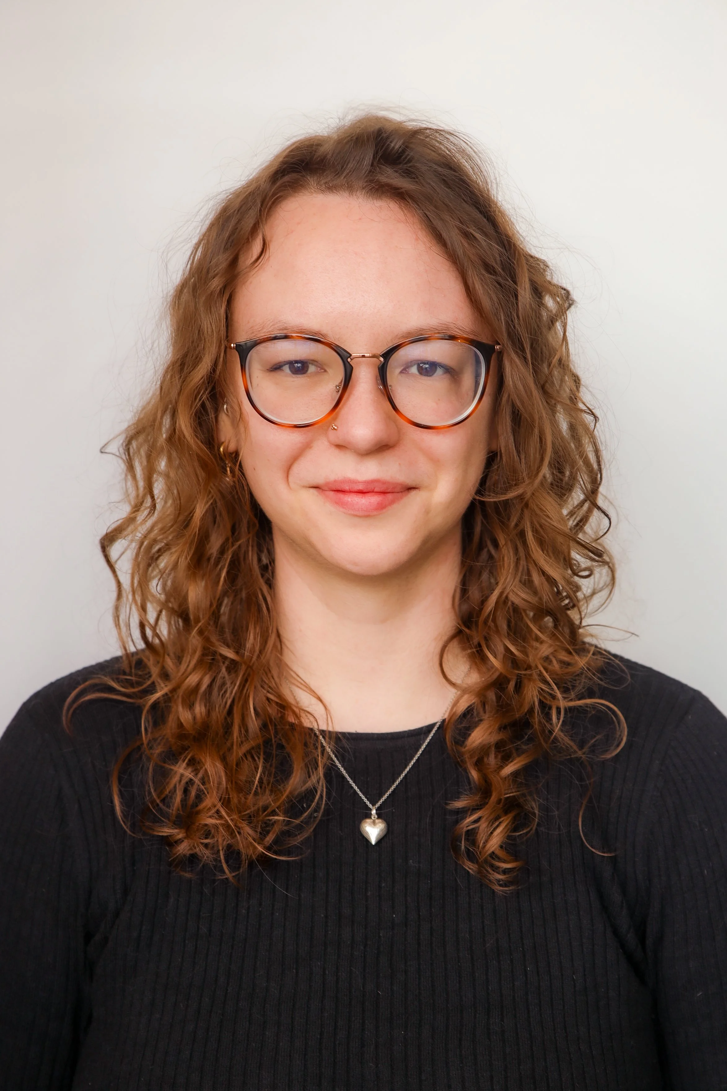 a white women with curly brown hair and brown glasses, wearing a black ribbed shirt and a silver heart necklace