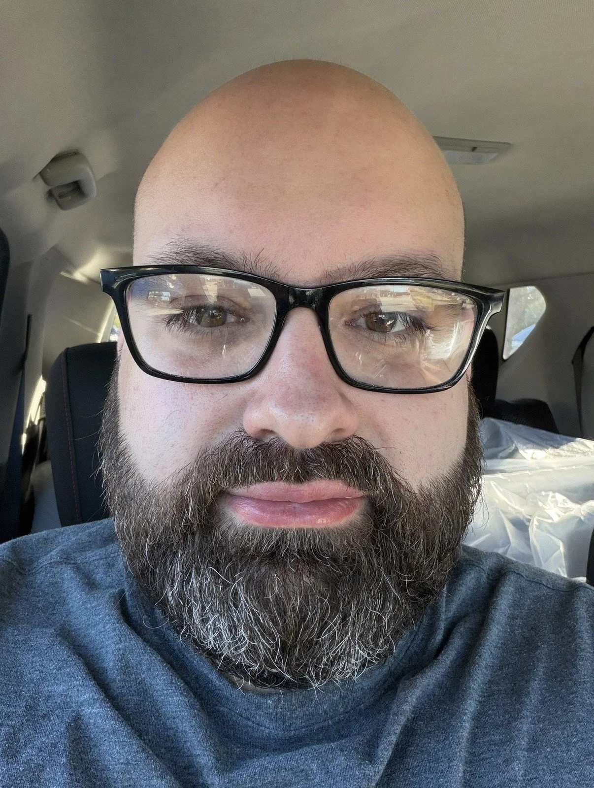 Close-up of a man with a beard, glasses, and a bald head, sitting in a vehicle.