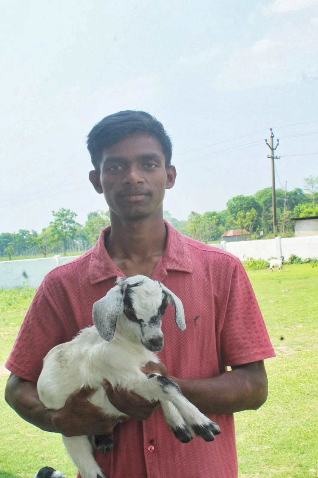 A young man holding a baby goat outdoors in a grassy area with trees and a fence in the background.