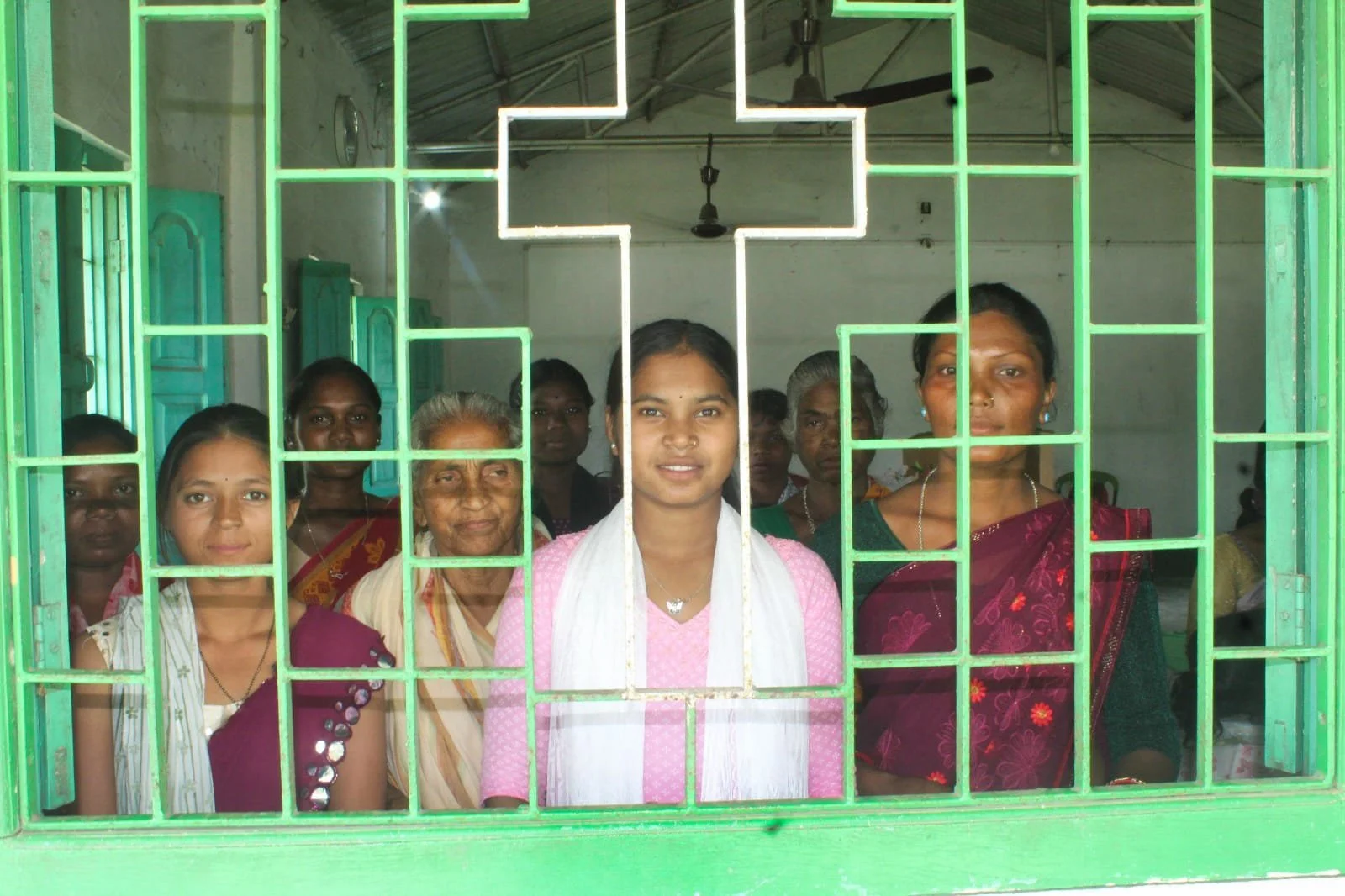 Group of women and girls standing behind a green metal grille with a cross symbol. Inside a room, they are looking through the grille toward the camera.