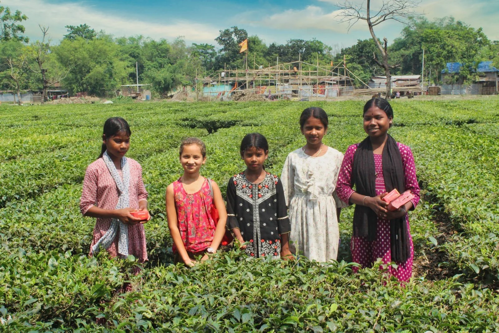 Five young girls standing in a lush green field with trees and a construction site in the background.