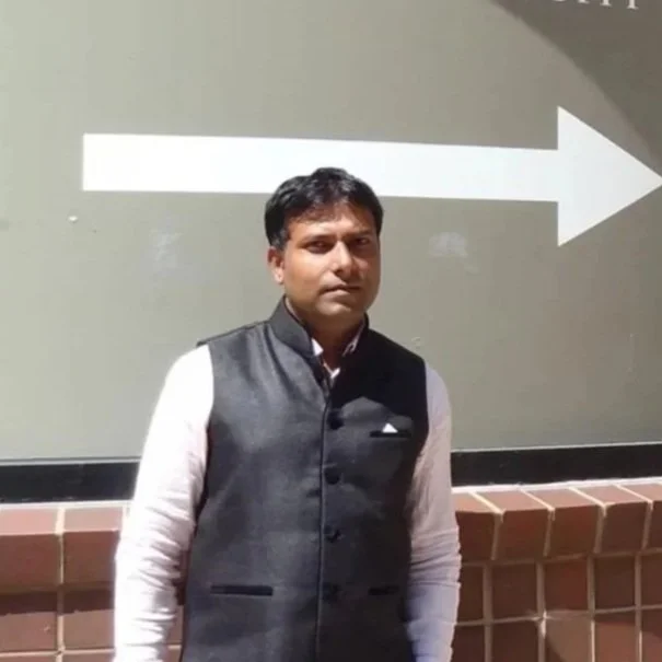 A man wearing a gray vest over a white shirt standing in front of a wall with a large white arrow pointing to the right.