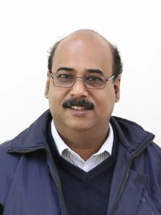 A man wearing glasses, a dark jacket, and a collared shirt, smiling against a plain white background.