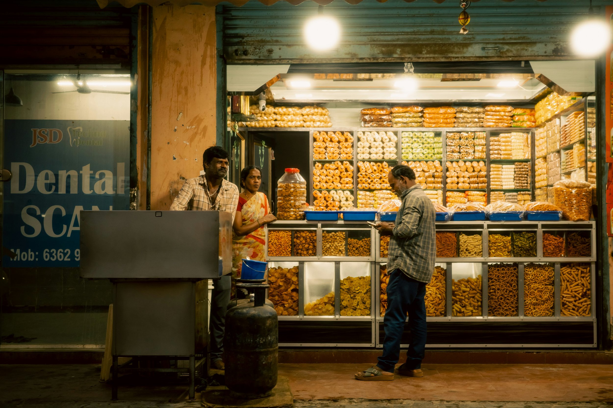 A street vendor shop selling various packaged snacks, with two men and one woman standing in front, illuminated at night.