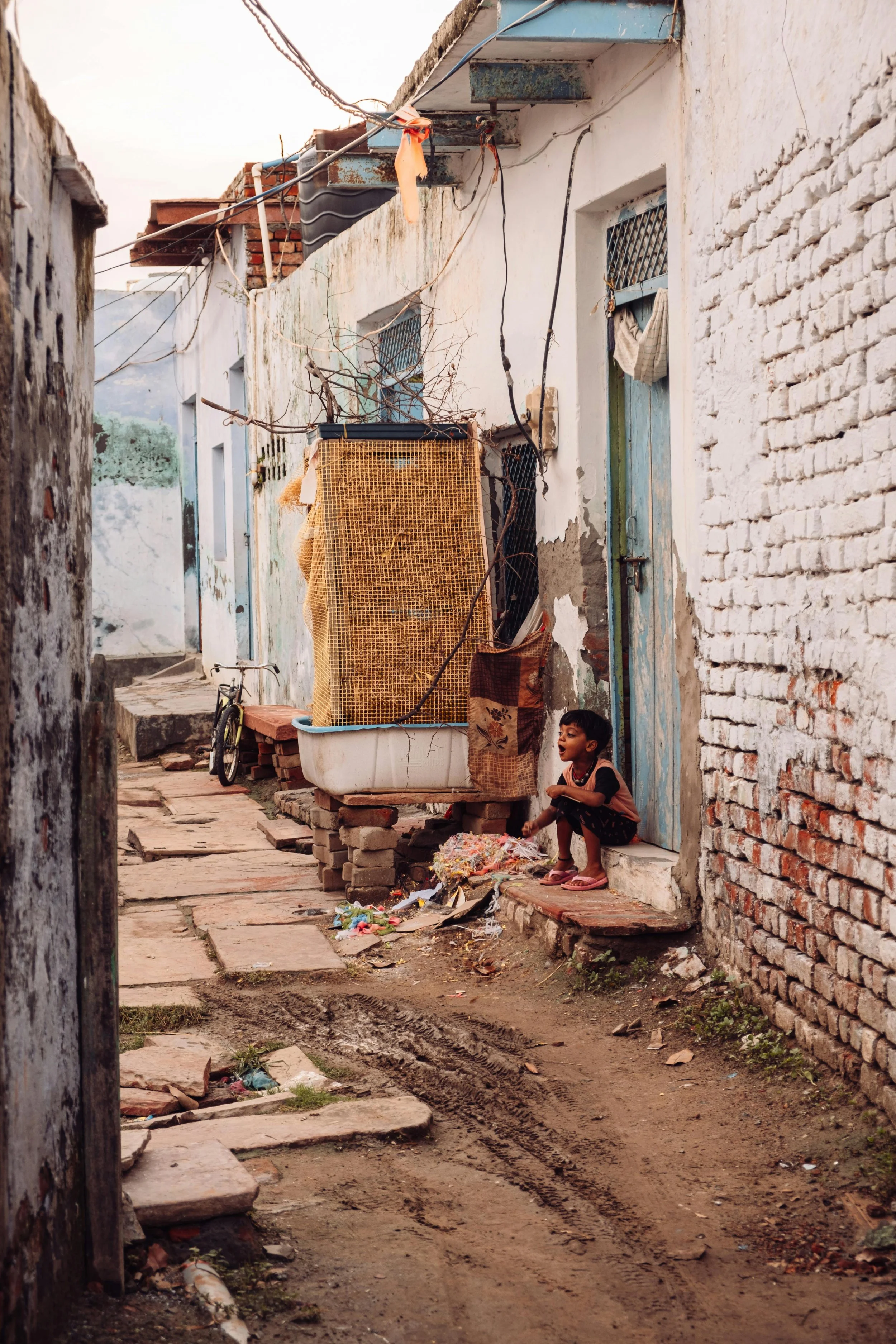A young boy squatting on a small step outside a worn house in a narrow alleyway with uneven, dirt and stone pavement, debris, and trash scattered around, with brick and painted concrete walls, a bicycle, and makeshift clothes drying lines overhead.