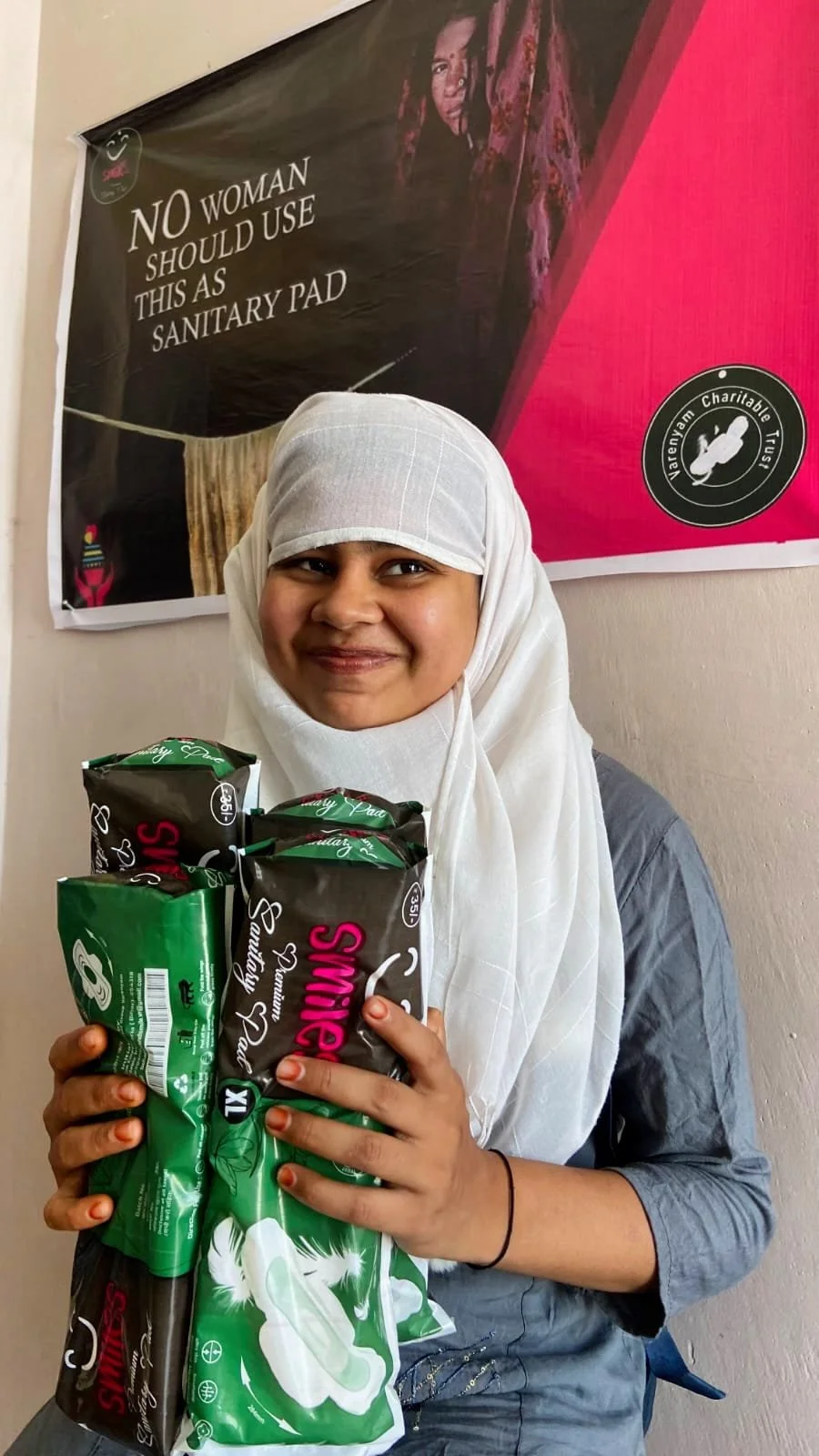 A smiling woman wearing a white headscarf and gray shirt holding several packages of sanitary pads. Behind her is a banner with text stating, 'NO WOMAN SHOULD USE THIS AS SANITARY PAD,' and a logo for Vatenyem Charitable Trust.