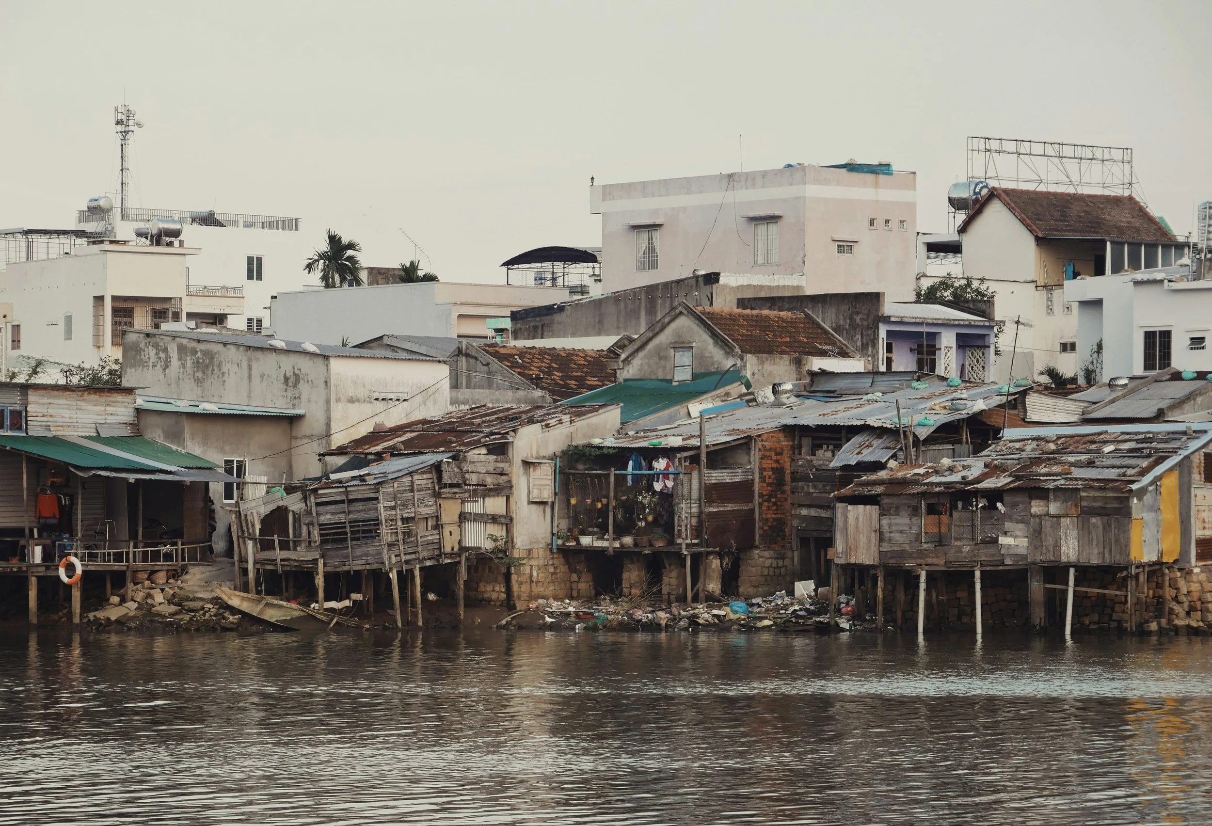 A riverside community with wooden stilt houses and modern buildings in the background under an overcast sky.