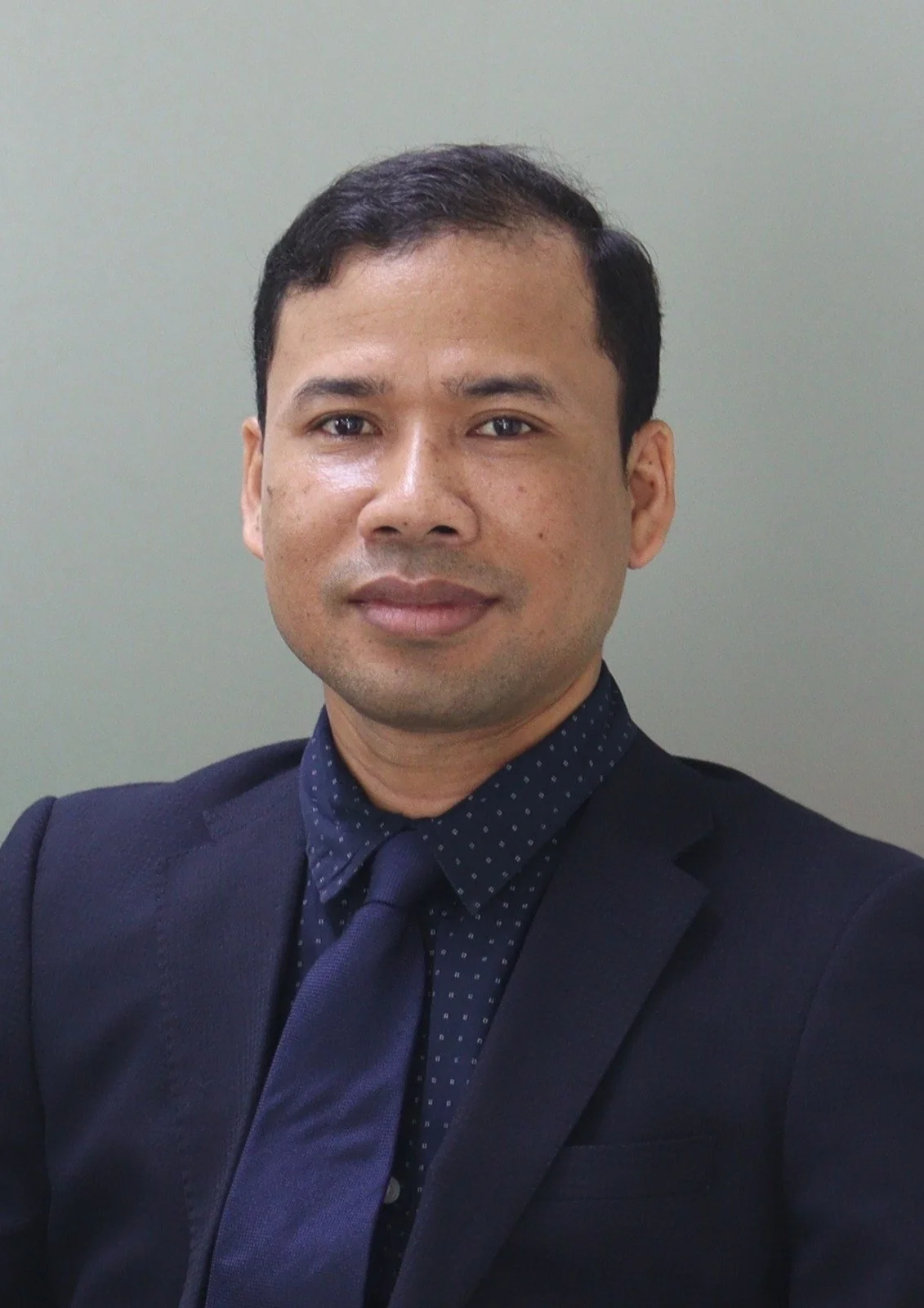 Portrait of a man in a dark suit, navy blue shirt with small white polka dots, and matching navy blue tie, standing against a plain light-colored background.